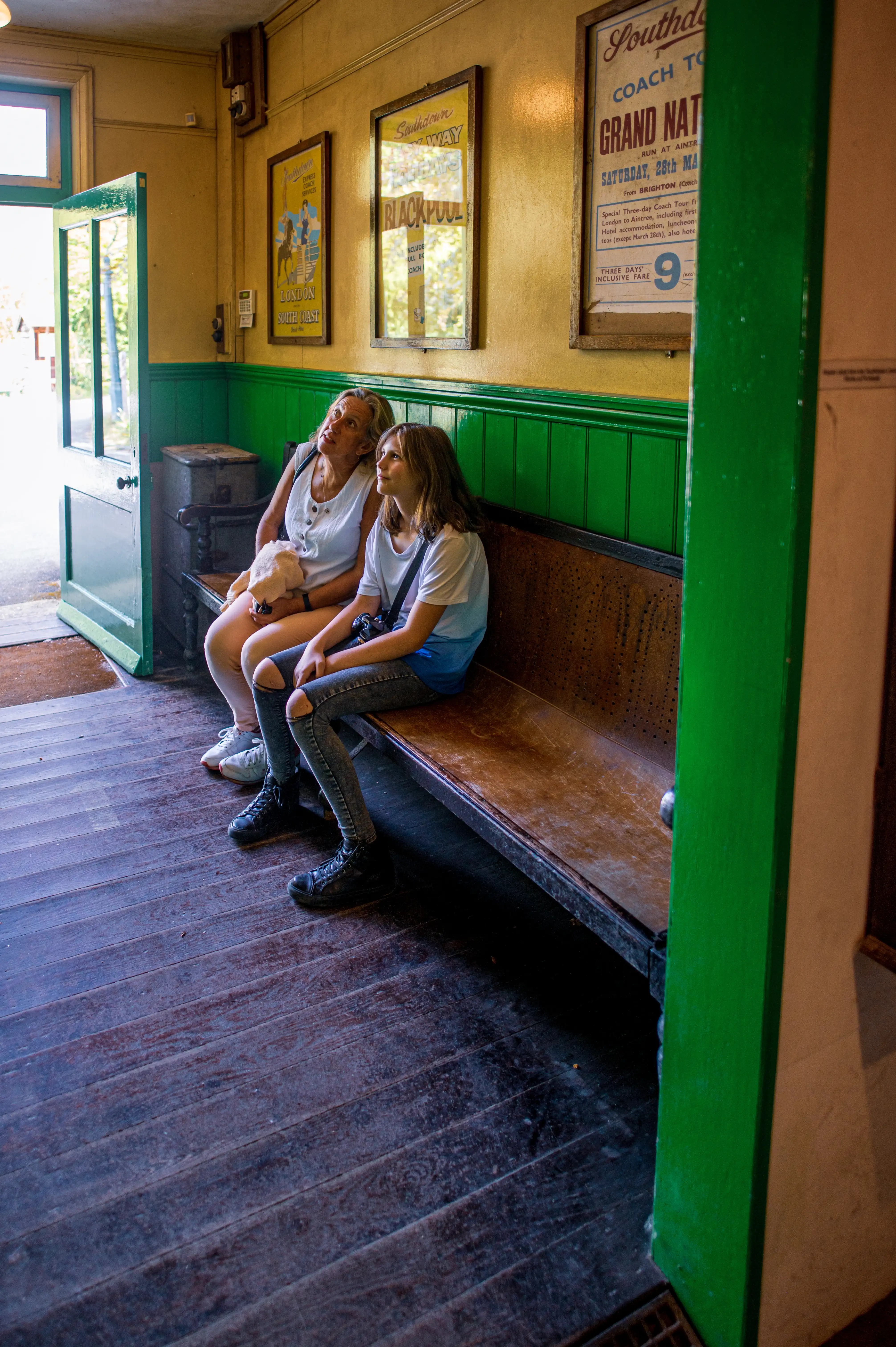 A woman and a teenage girl sit on a wooden bench in a vintage train station waiting room, looking upwards.