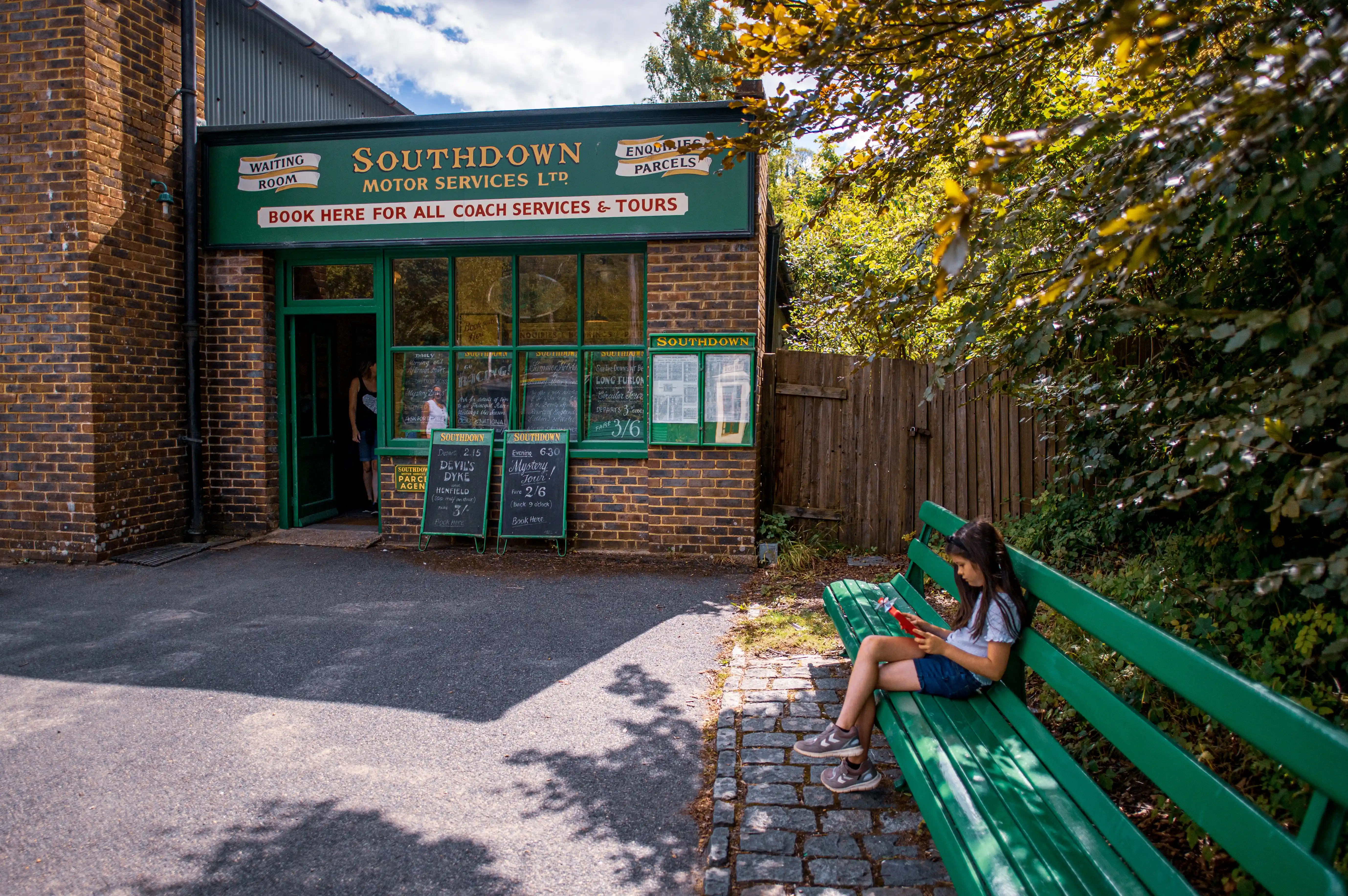 Small brick Southdown Motor Services booking office with green trim, chalkboard notices and a child on a bench.