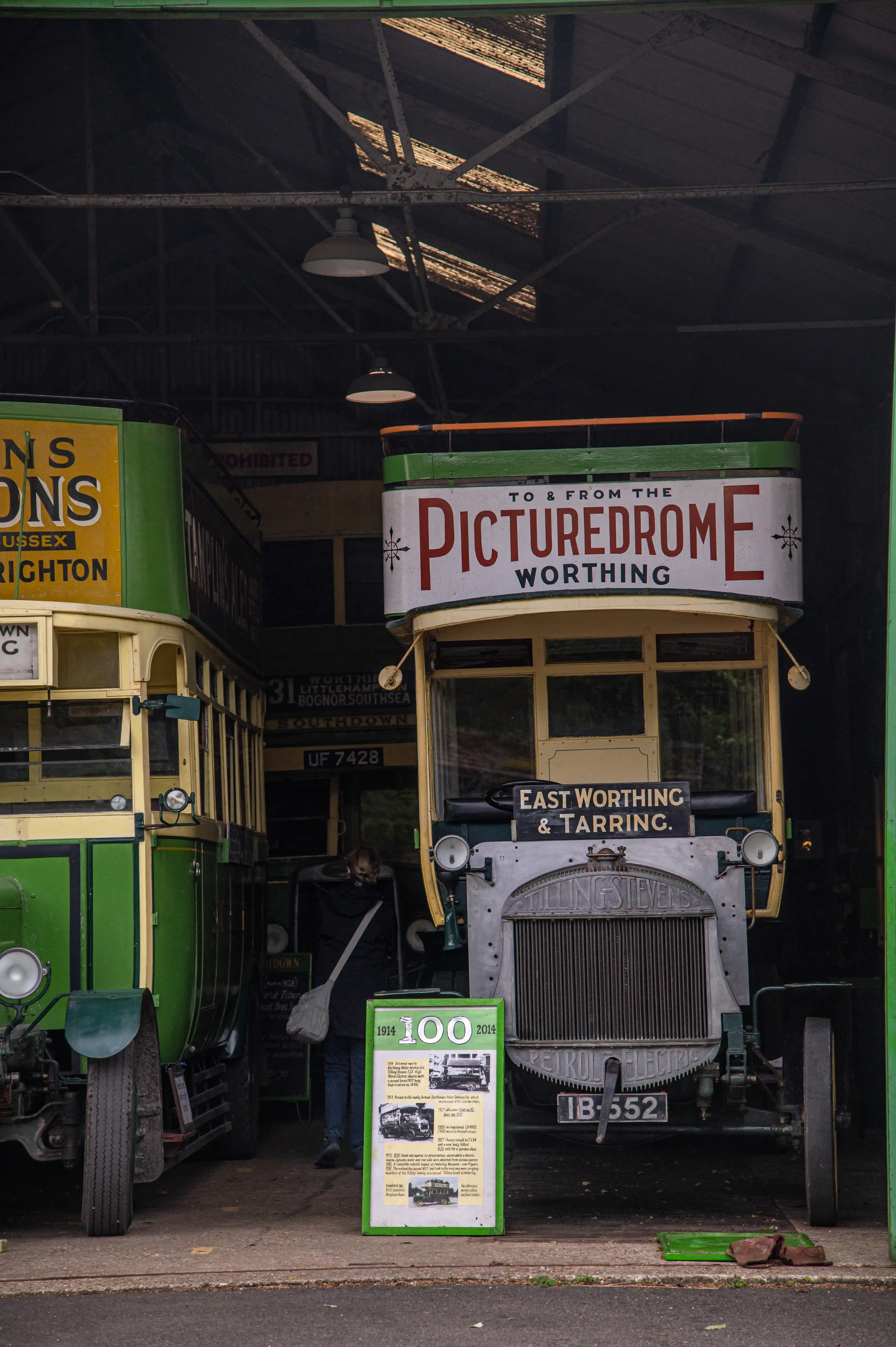 A view inside a shed showing the front of two vintage buses. The bus on the right has a sign that reads 'PICTUREDROME WORTHING' and 'EAST WORTHING & TARRING.' A green display board with historical information and images is in front of the bus.