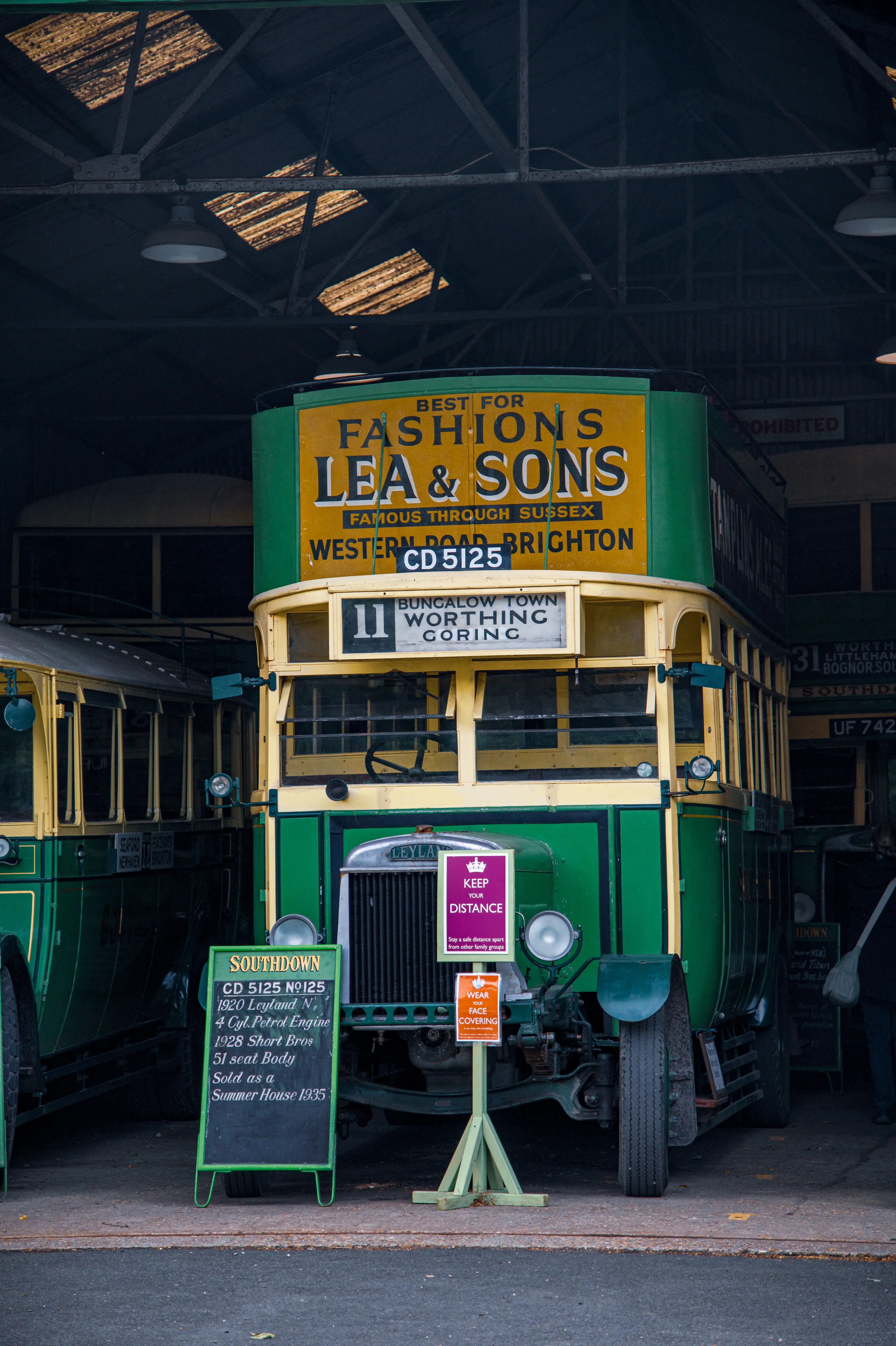 A vintage green and yellow Southdown double-decker bus with the registration CD 5125 is parked in a shed. A blackboard in front of the bus provides its specifications, and route destination signs are visible on the bus.