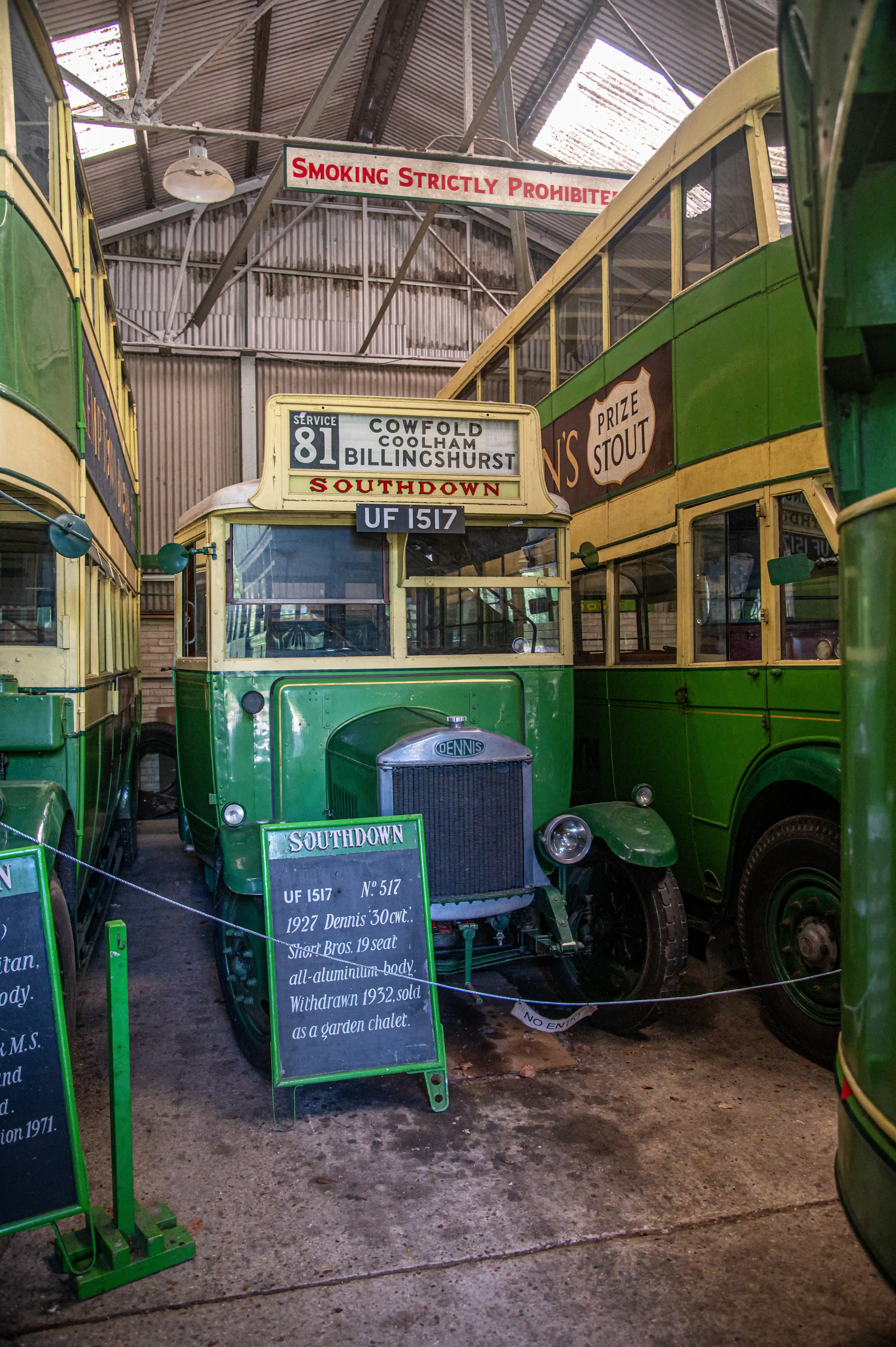 Interior view of a shed housing several vintage green and cream Southdown buses. The foreground bus, a 1927 Dennis with registration UF 1517, is central, with a route board indicating Service 81 to Cowfold, Coolham, and Billingshurst. A smaller blackboard beside it provides details about the bus, noting it was withdrawn in 1932 and later sold as a garden chalet. The metallic grille of the bus is branded 'DENNIS'. A 'Prize Stout' advertisement is visible on the side of a bus to the right. A red sign overhead warns 'SMOKING STRICTLY PROHIBITED'.