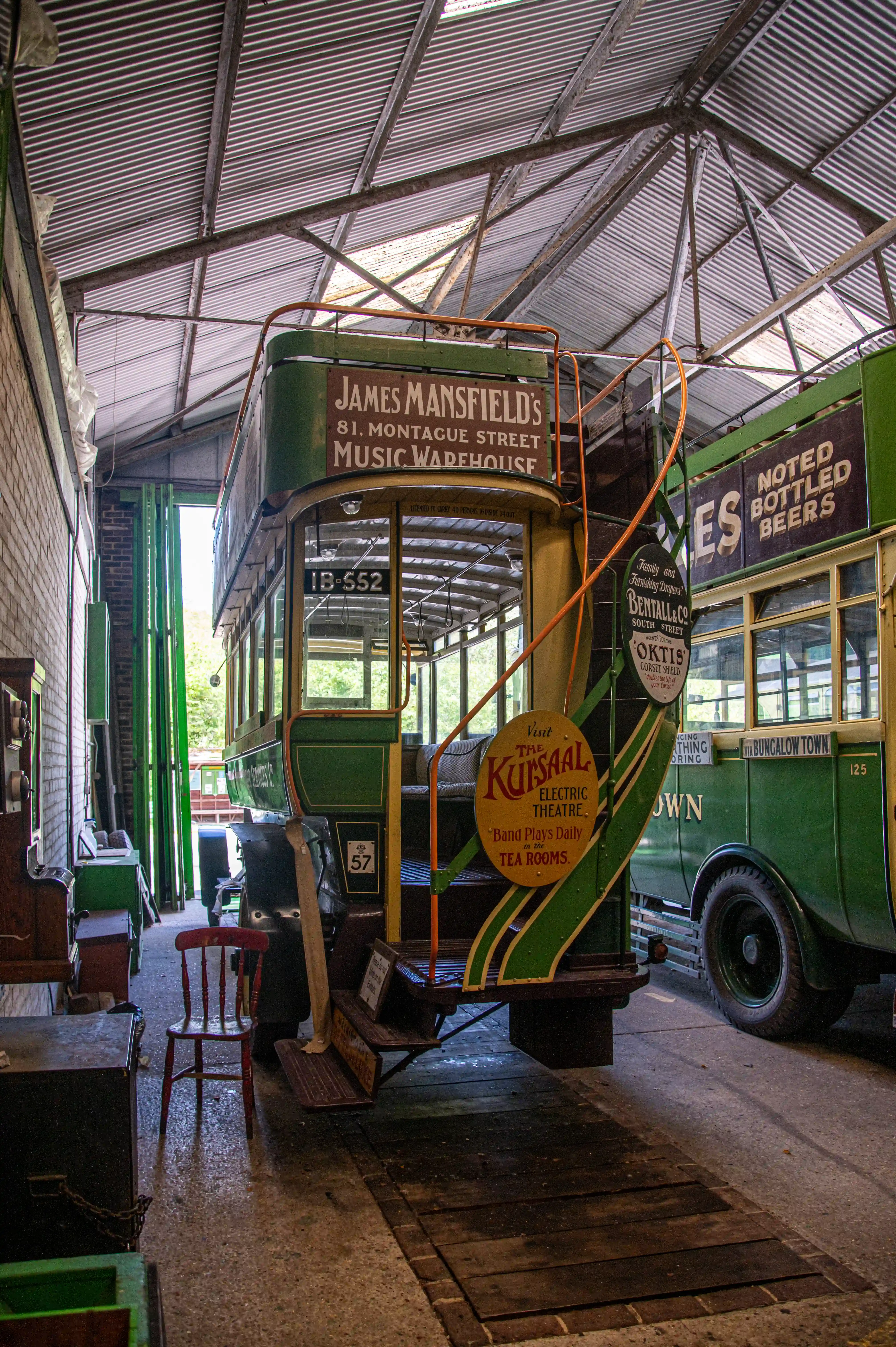 A photograph showing two vintage green and cream trams inside a barn with a corrugated iron roof. The tram on the left has signage advertising a music warehouse and a theatre, while the tram on the right has advertisements for bottled beers and a place called Bungalow Town.