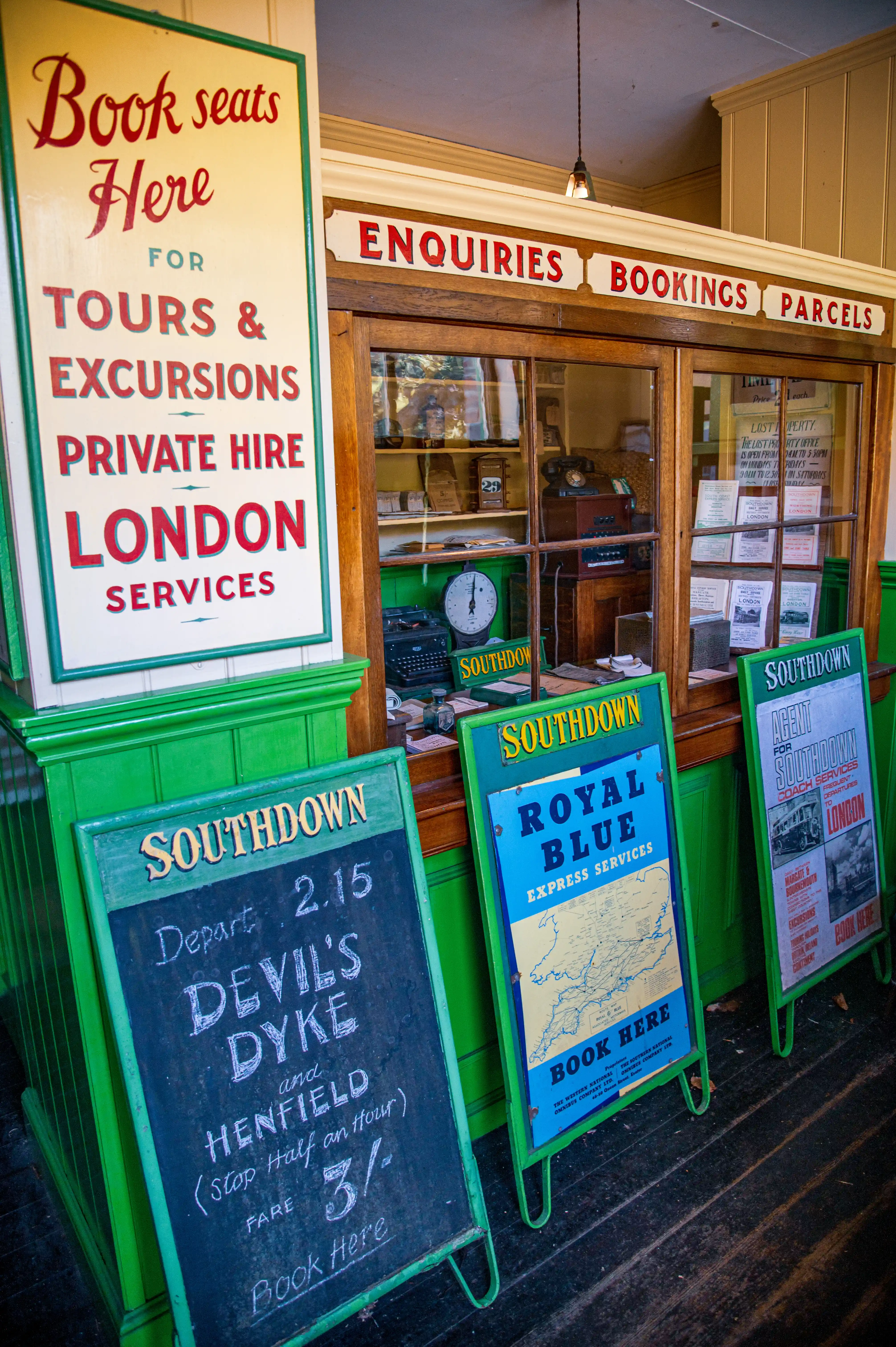 A vintage Southdown bus ticket office with a sign reading 'Book seats Here' and three A-frame boards advertising bus routes and fares, including 'Devil's Dyke & Henfield' and 'Royal Blue Express Services.'