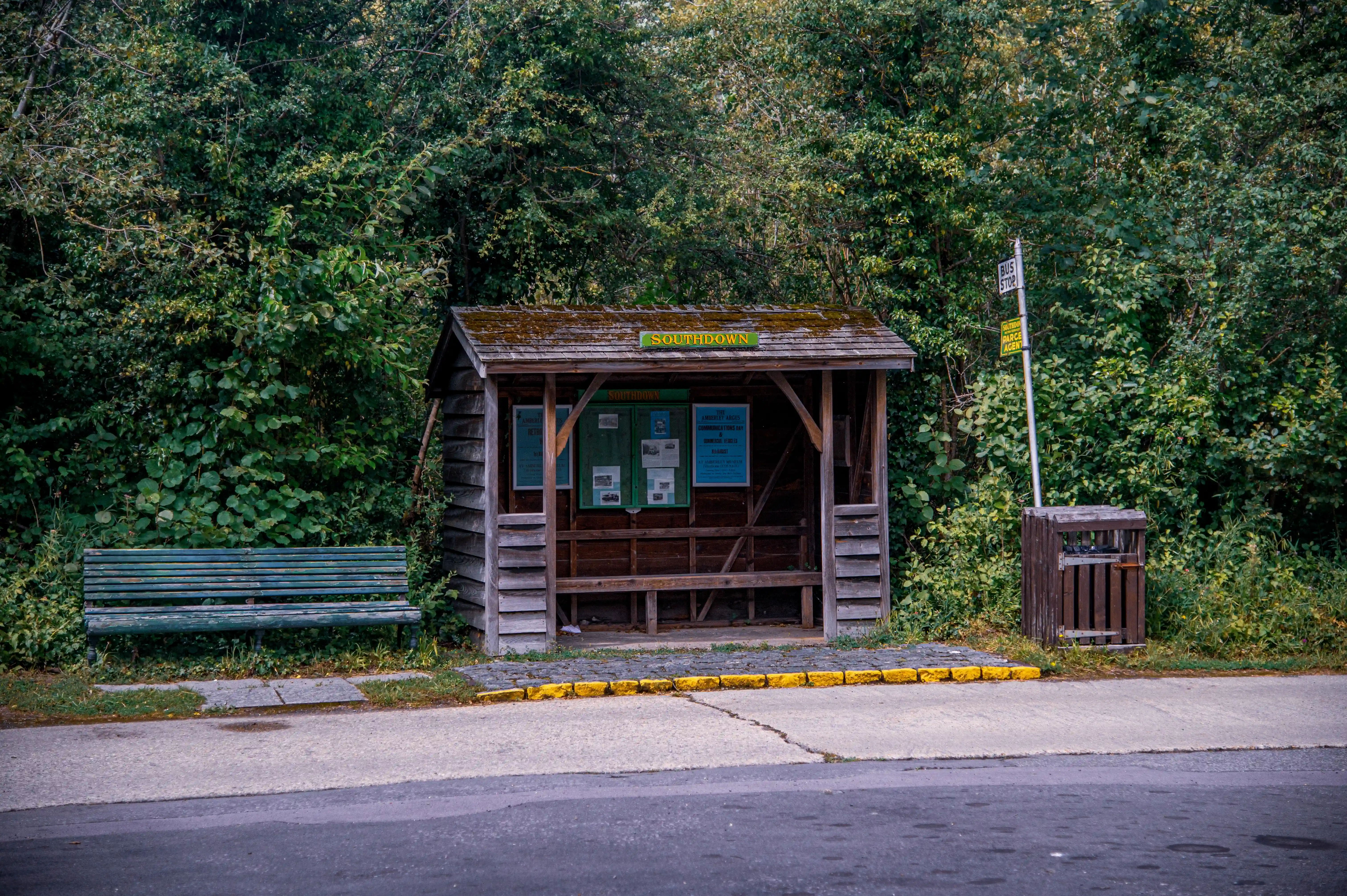 A rustic wooden bus stop shelter with a green sign reading 'SOUTHDOWN' is situated next to a green bench and a wooden bin, with a dense green forest in the background. A bus stop sign is also visible.