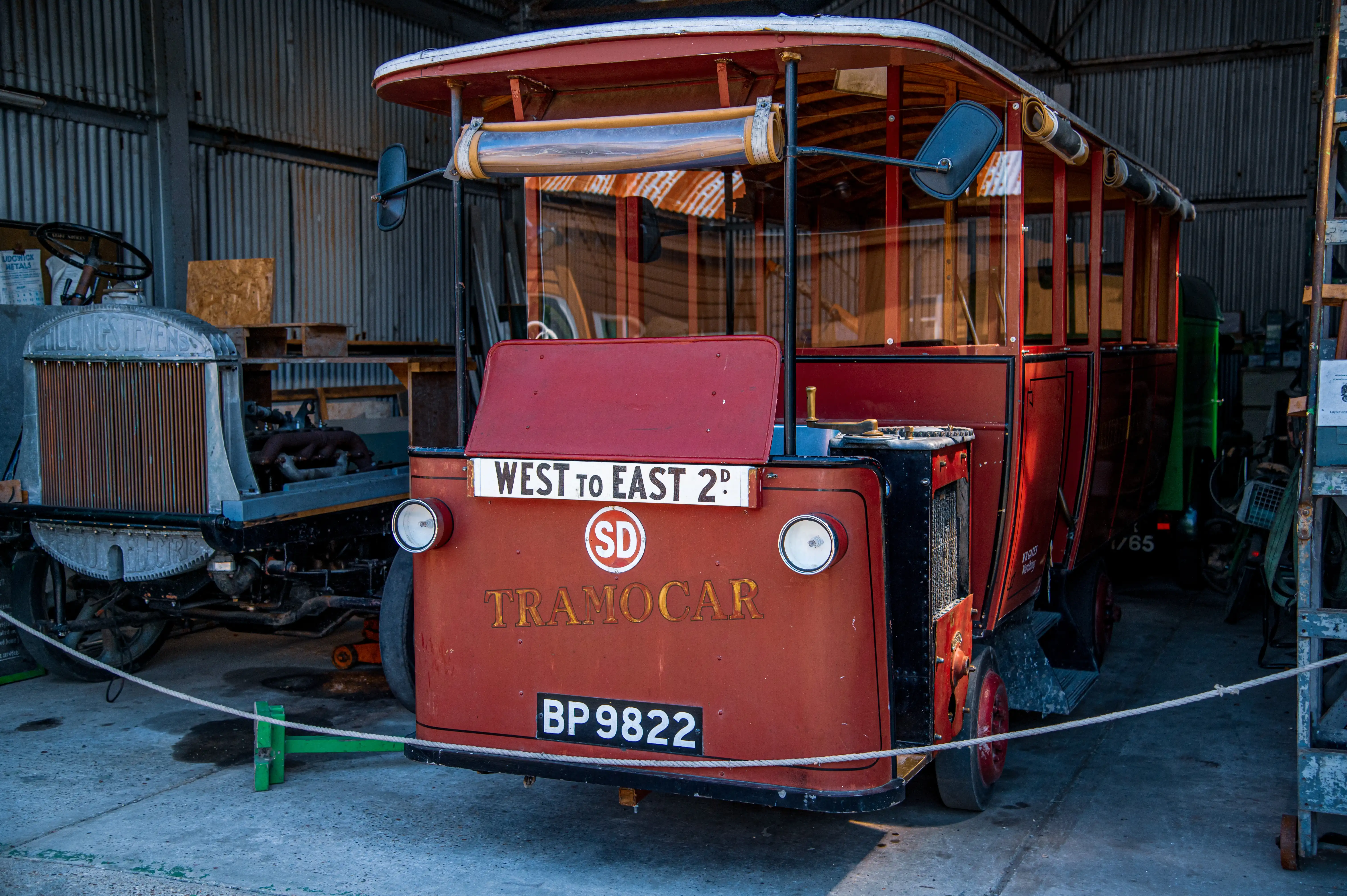 A close-up front view of a vintage red tramocar with wooden accents and a destination sign reading 'WEST TO EAST 2D'. The vehicle has a registration plate 'BP 9822'. Another older vehicle is partially visible to the left.