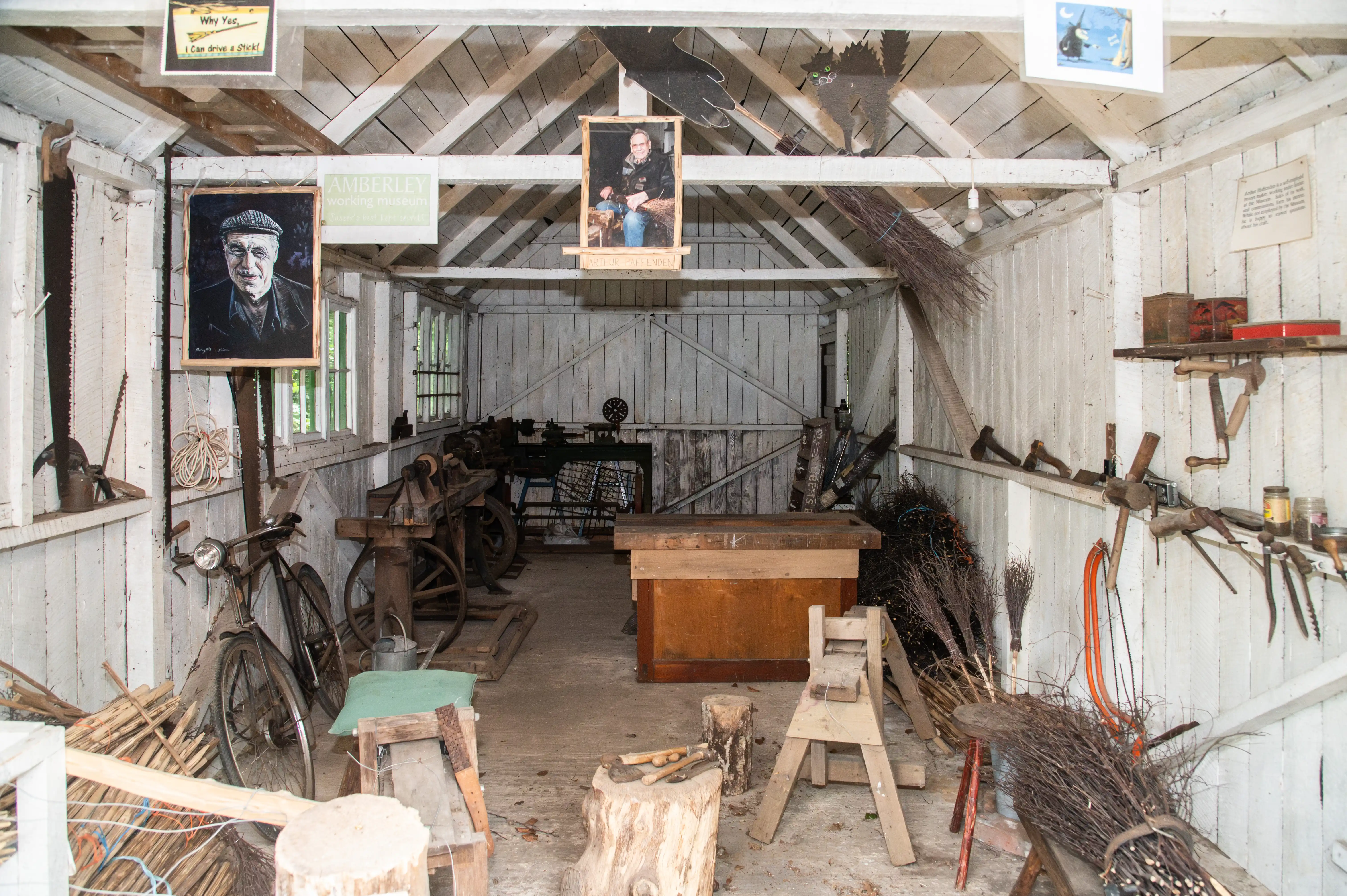 Interior view of a rustic wooden workshop filled with tools and materials for broom making, including lathes, bundles of twigs, and portraits on the walls.
