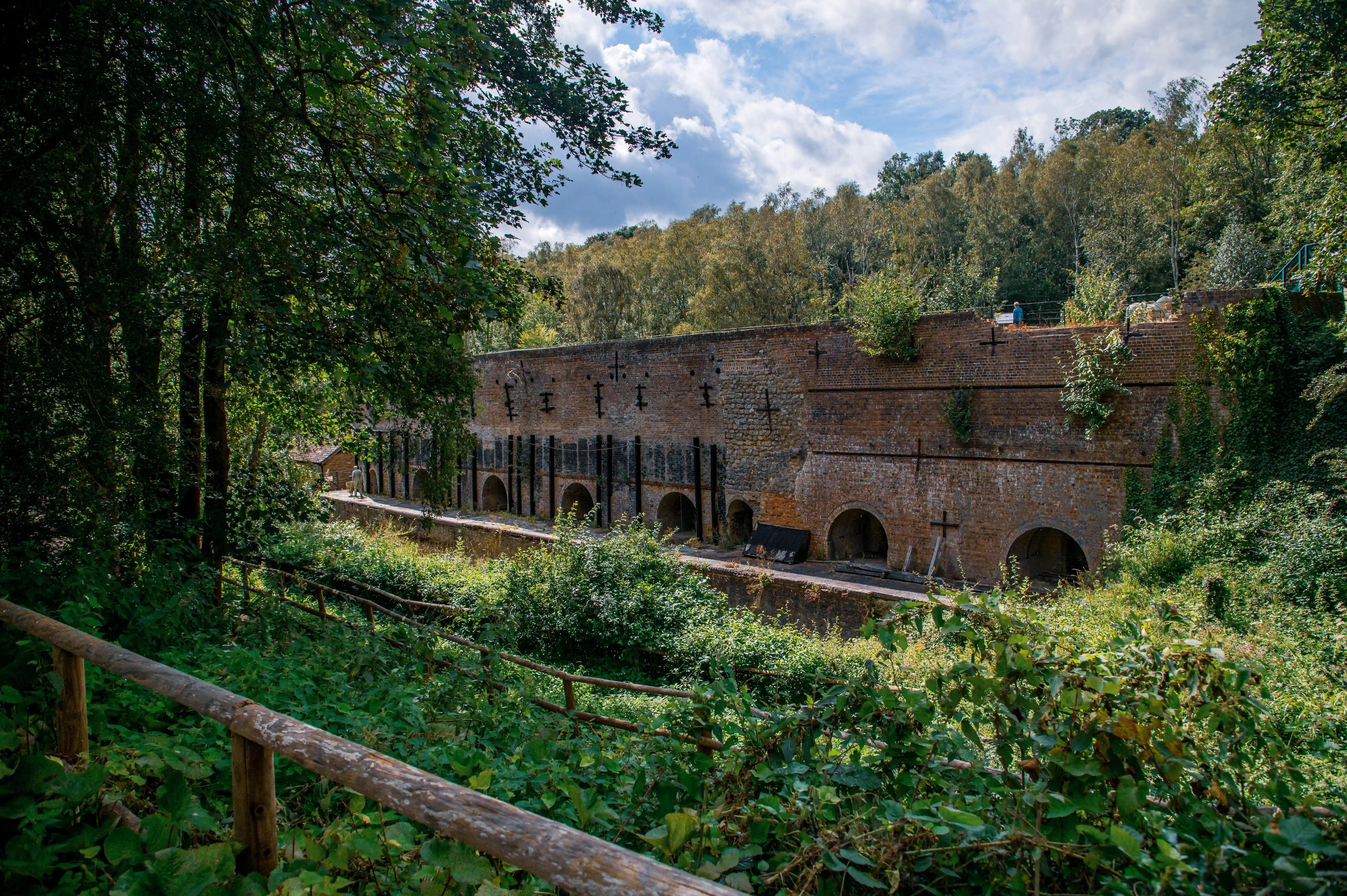Old brick industrial structure with arched openings and metal supports, surrounded by dense greenery and a wooden fence.