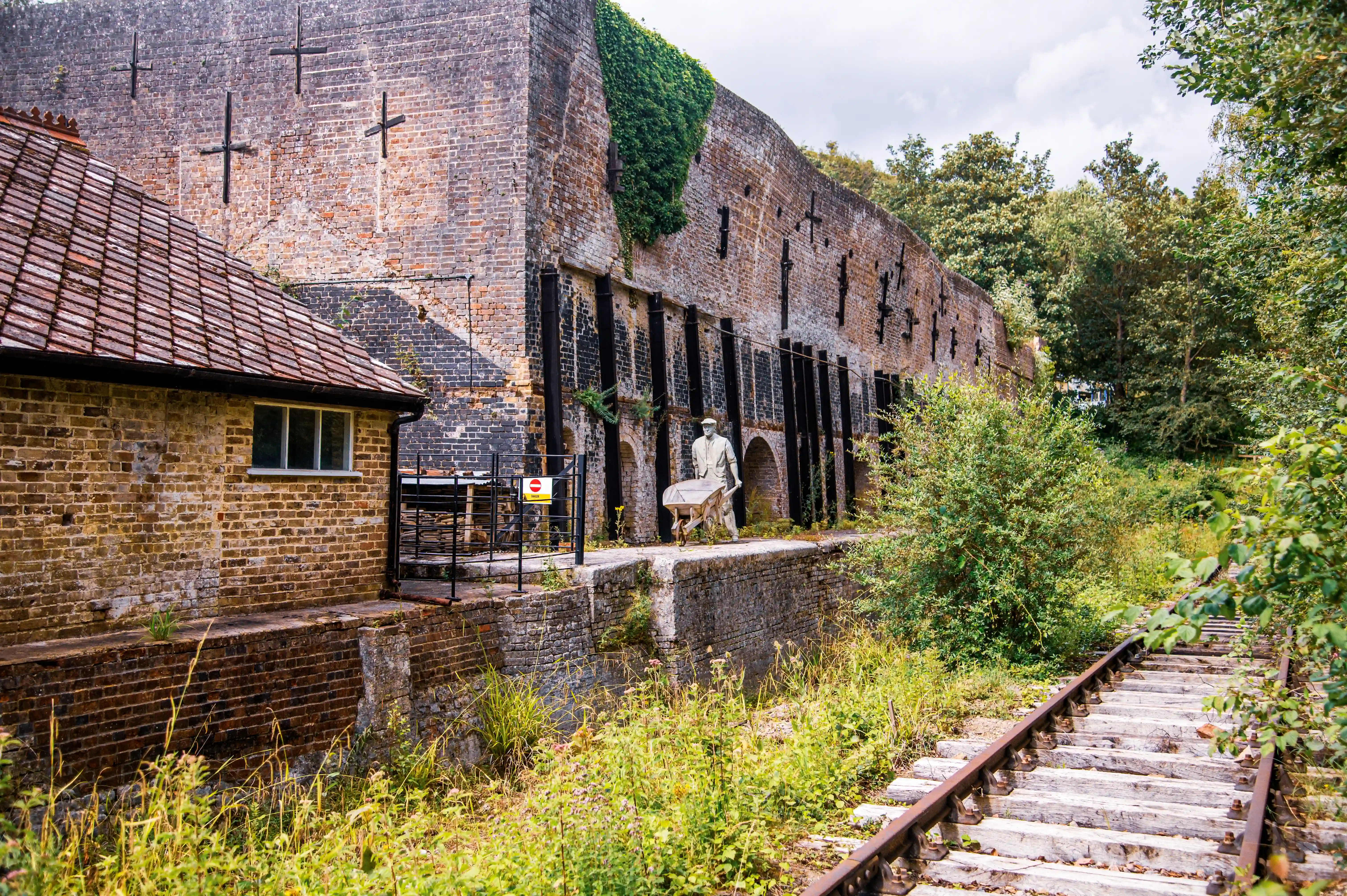 A statue of a man pushing a wheelbarrow stands in front of a large, old brick industrial building. Railway tracks run in front of the building, bordered by overgrown grass and bushes.
