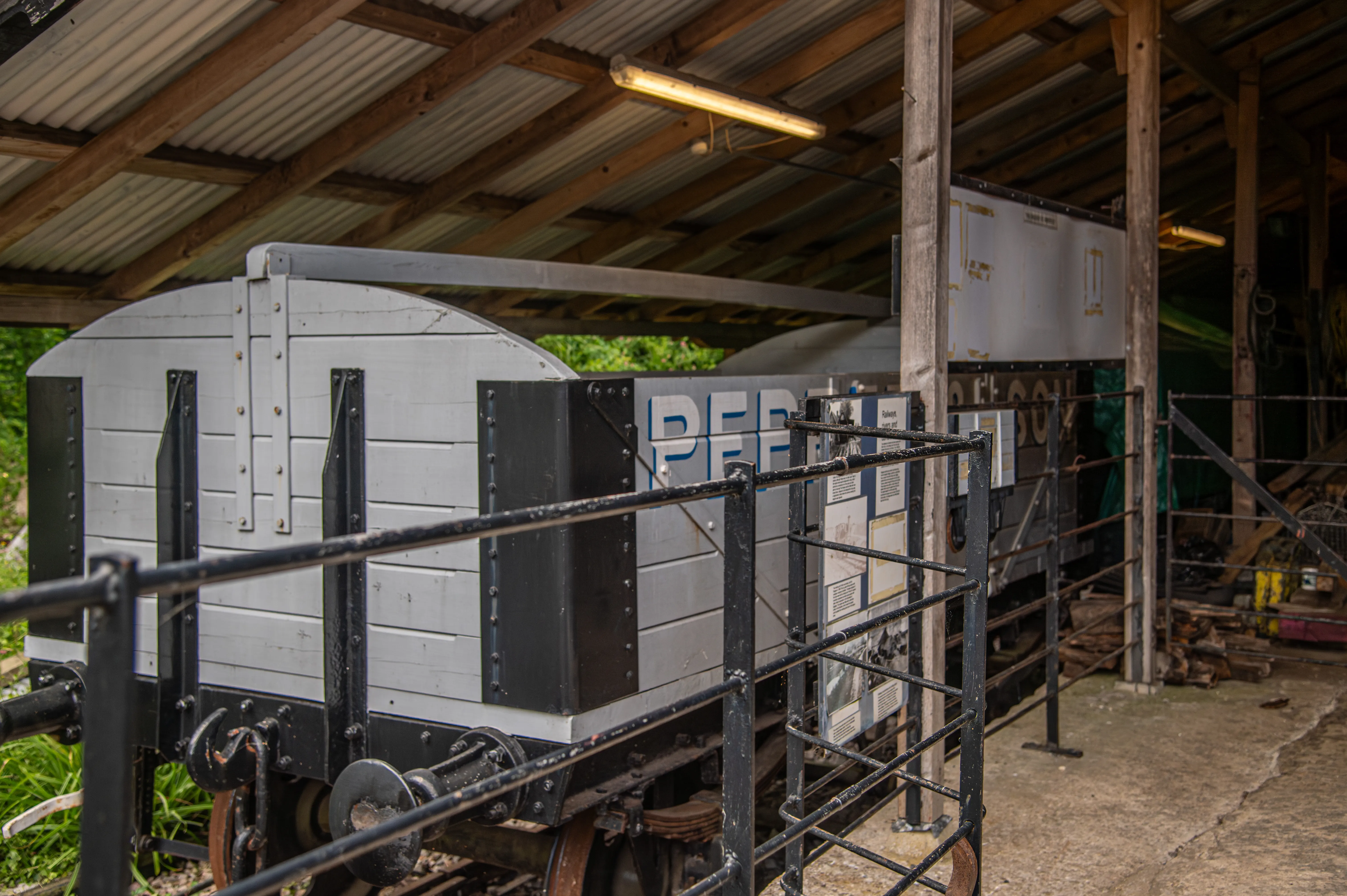 Grey railway wagon with black metal reinforcements displayed under a covered shed with safety railing and museum information boards.