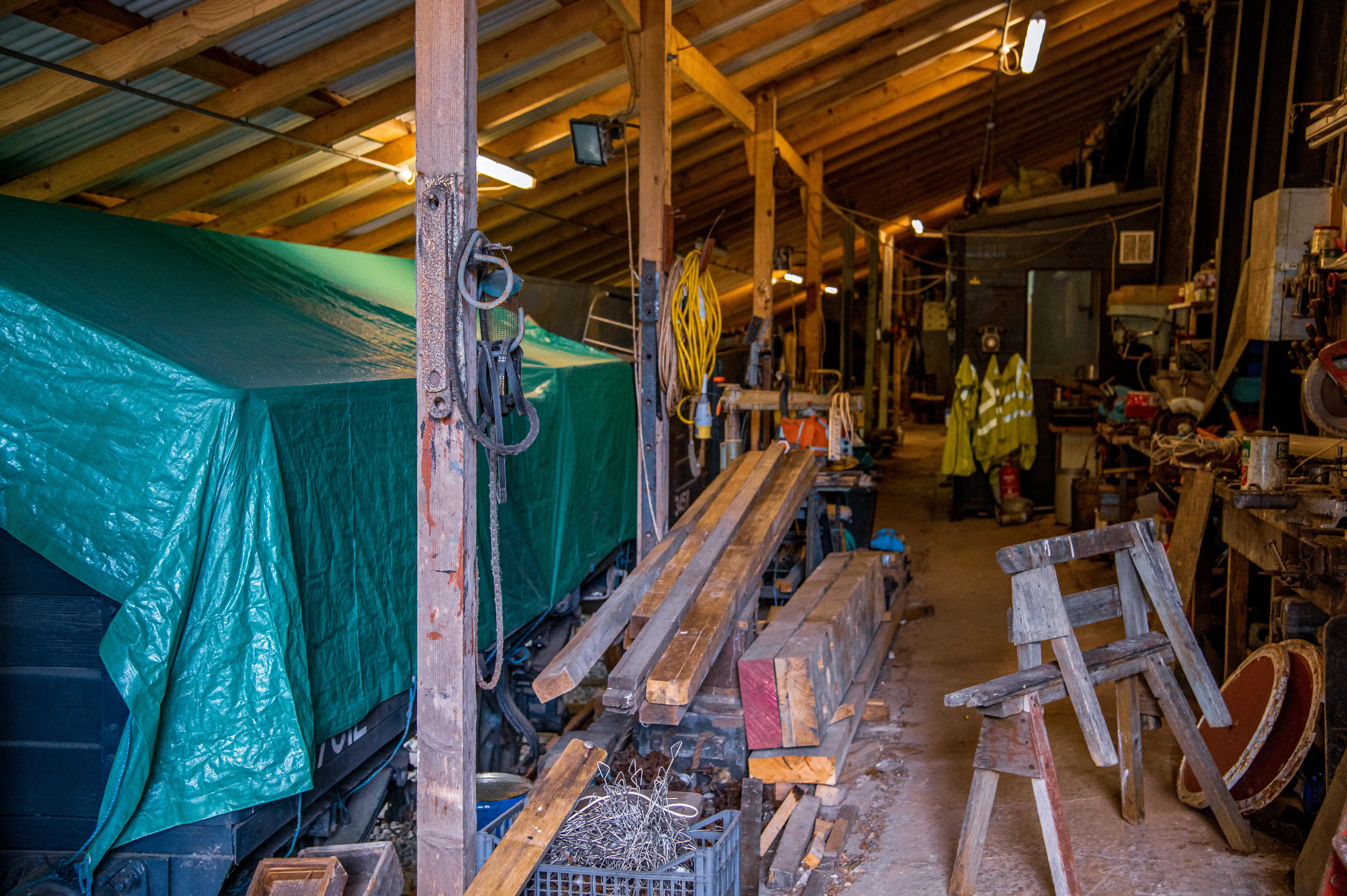Interior of a cluttered workshop with wooden beams, tools, lumber, and a green covered object.