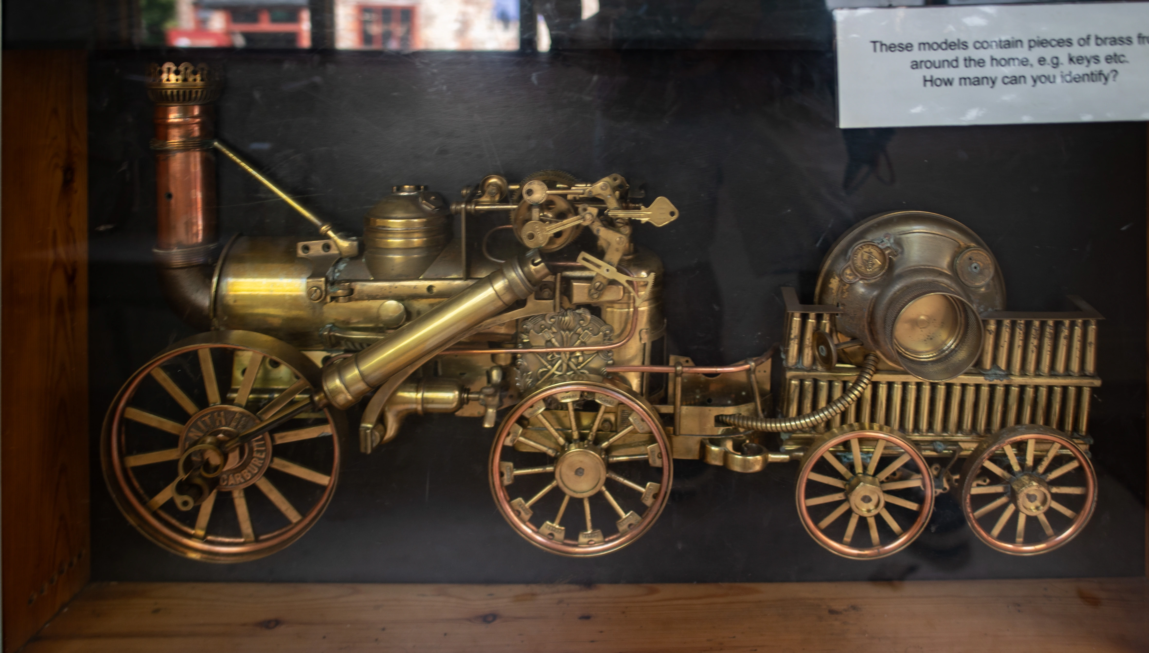 A close-up, side view of a brass model steam locomotive, showcasing its intricate machinery, wheels, and tender. A sign behind the model asks viewers to identify brass parts used.