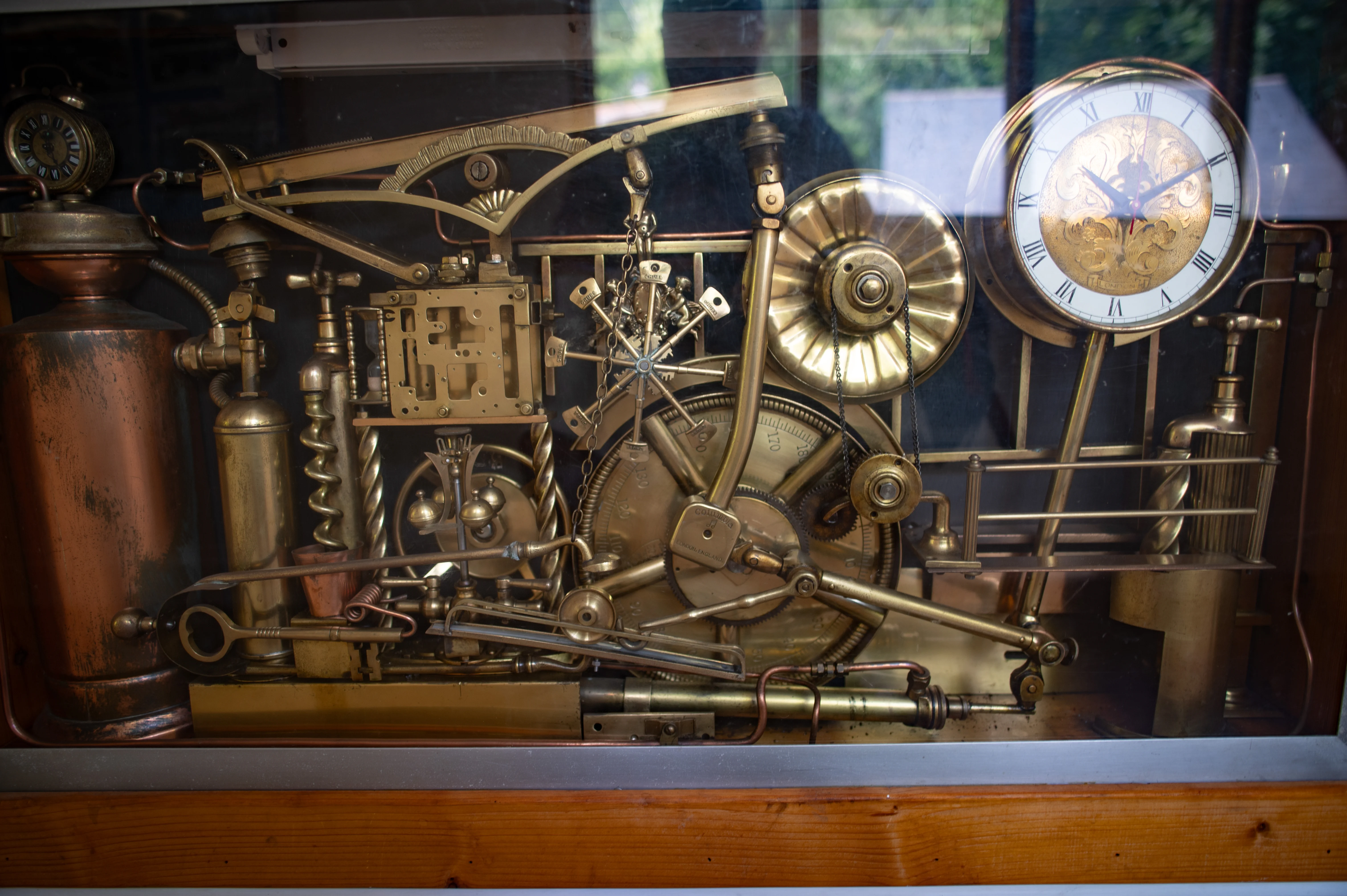 A complex, multi-component brass and copper clockwork mechanism displayed behind glass. Key features include gears, levers, chains, a pendulum, and a clock face with Roman numerals.