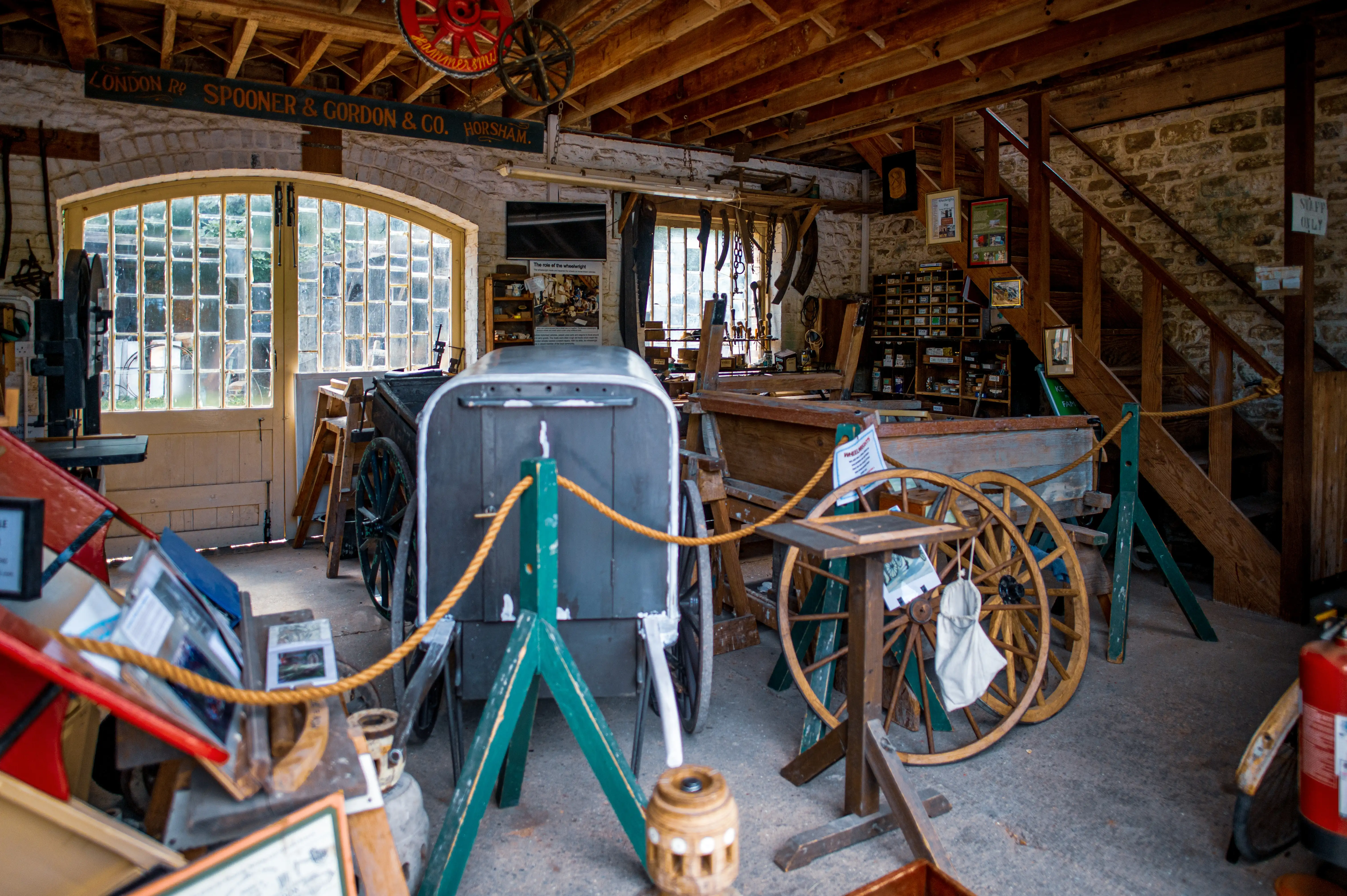 Interior view of a rustic wheelwright's workshop, filled with antique carts, wooden wheels, tools, and display boards, illuminated by natural light from large windows.