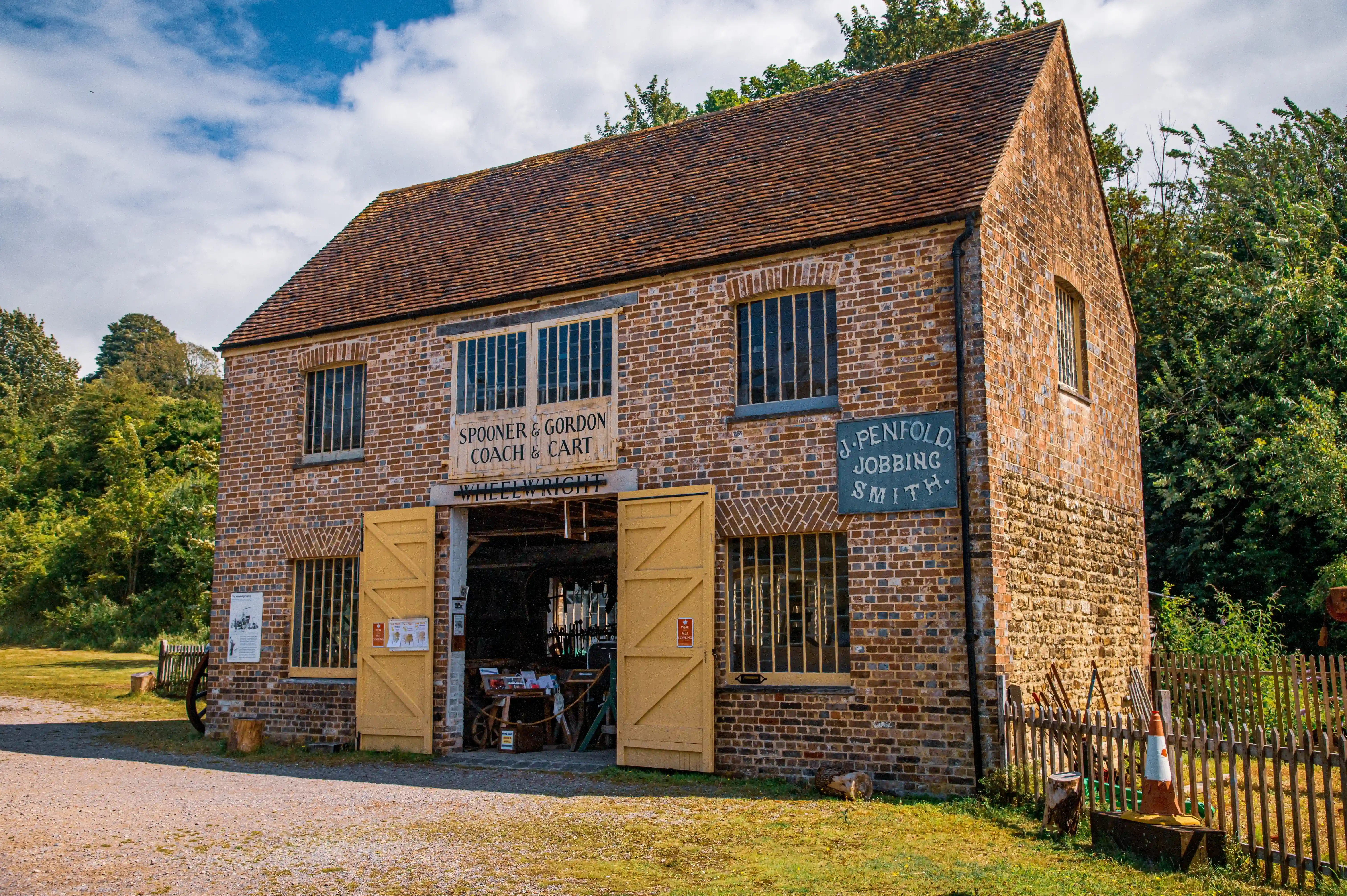 A historic brick building with a red tiled roof, featuring large yellow double doors that are open, revealing the interior of a workshop. Two signs are visible on the building's facade: one reads 'SPOONER & GORDON COACH & CART WHEELWRIGHT' and another reads 'J. PENFOLD JOBBING SMITH.' The workshop appears to be stocked with tools. The building is set within a grassy area with trees and a wooden fence.