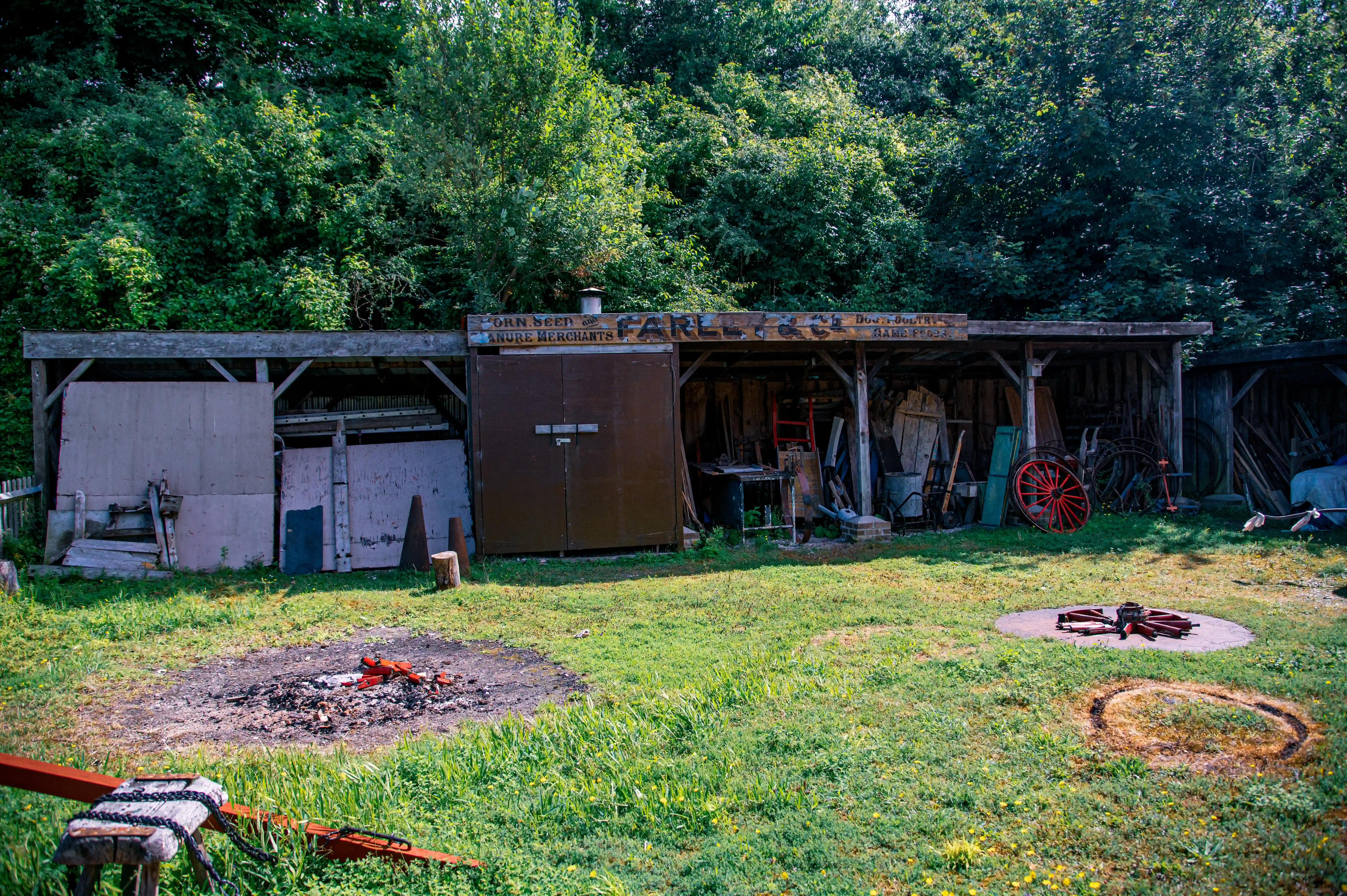An old wooden shed filled with various tools and objects, including antique red and black wagon wheels, stands in a grassy yard with a fire pit.