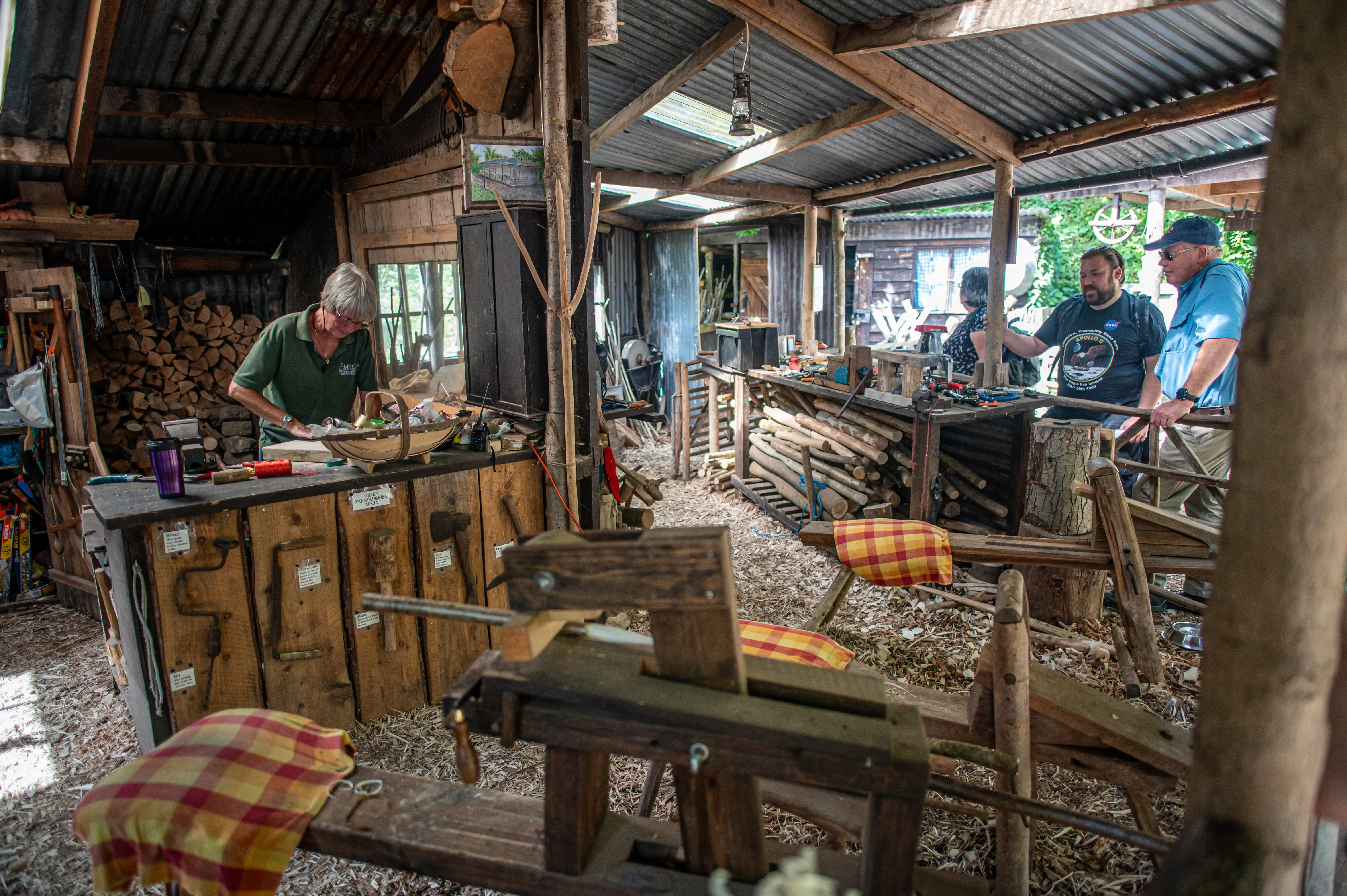 Three men are in a rustic wooden shed filled with tools and wood. One man in a green shirt is working at a workbench, while two others stand nearby, conversing.