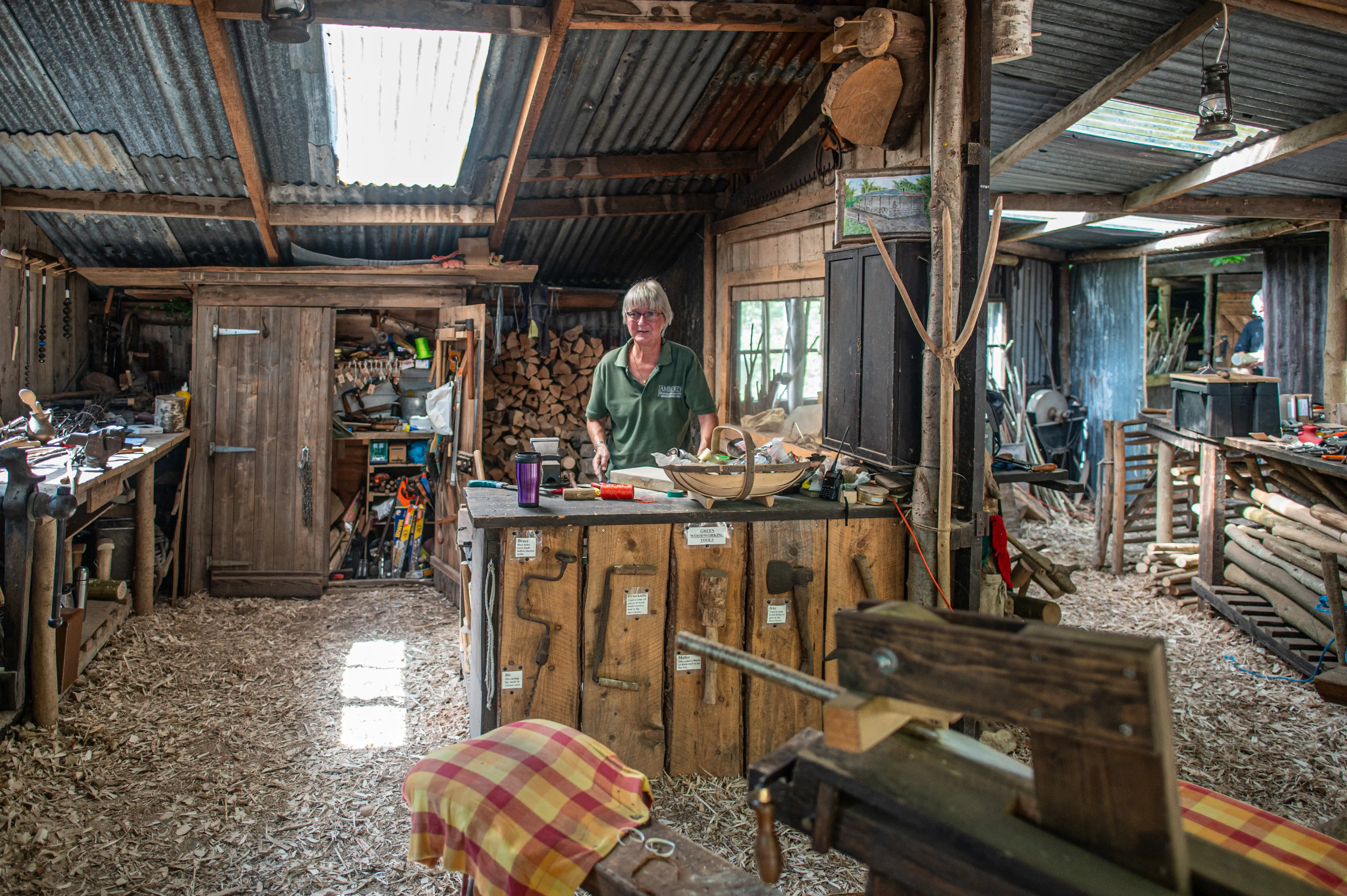 A woman in a green polo shirt stands in a cluttered, rustic workshop filled with woodworking tools, wood shavings on the floor, and stacks of logs.