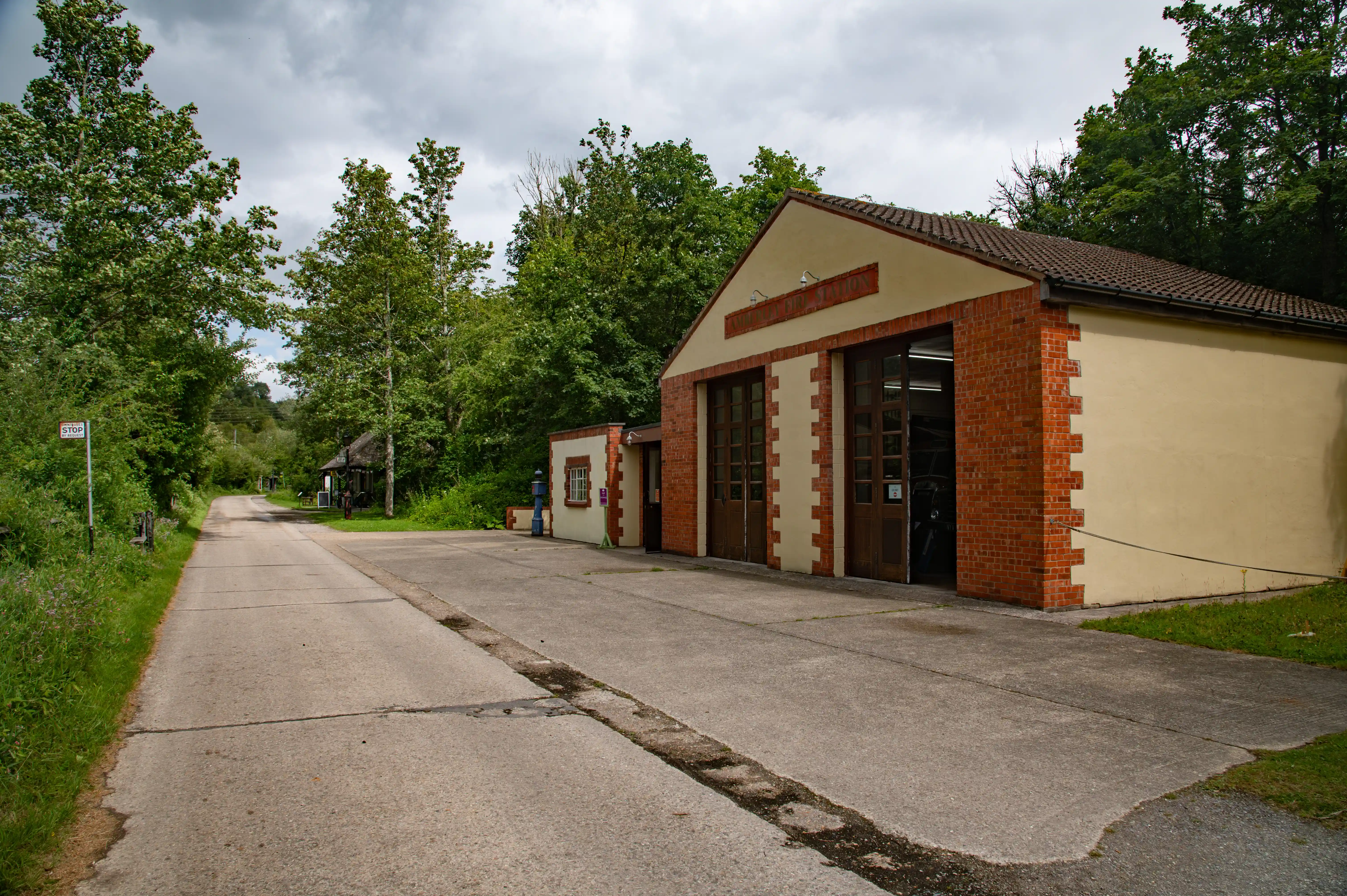 Exterior view of a charming brick and cream-coloured fire station building with large wooden doors, situated on a quiet rural road surrounded by dense trees.