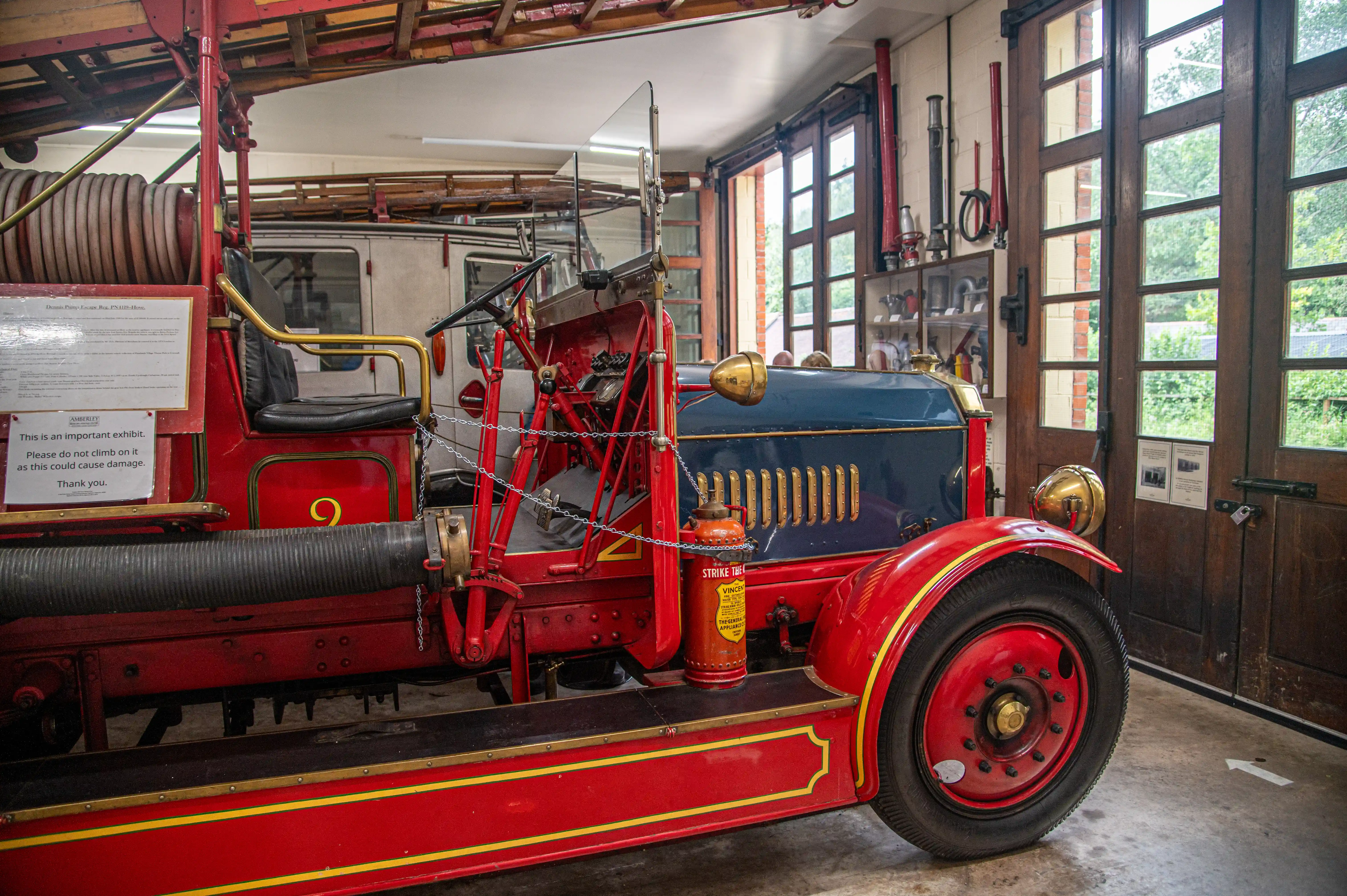 A vintage red and blue fire engine with the number 9 prominently displayed, sits in a garage. Brass headlights and accents gleam on the vehicle, which has a large hose wound around a reel on the side. Other firefighting equipment and large wooden doors with glass panes are visible in the background.