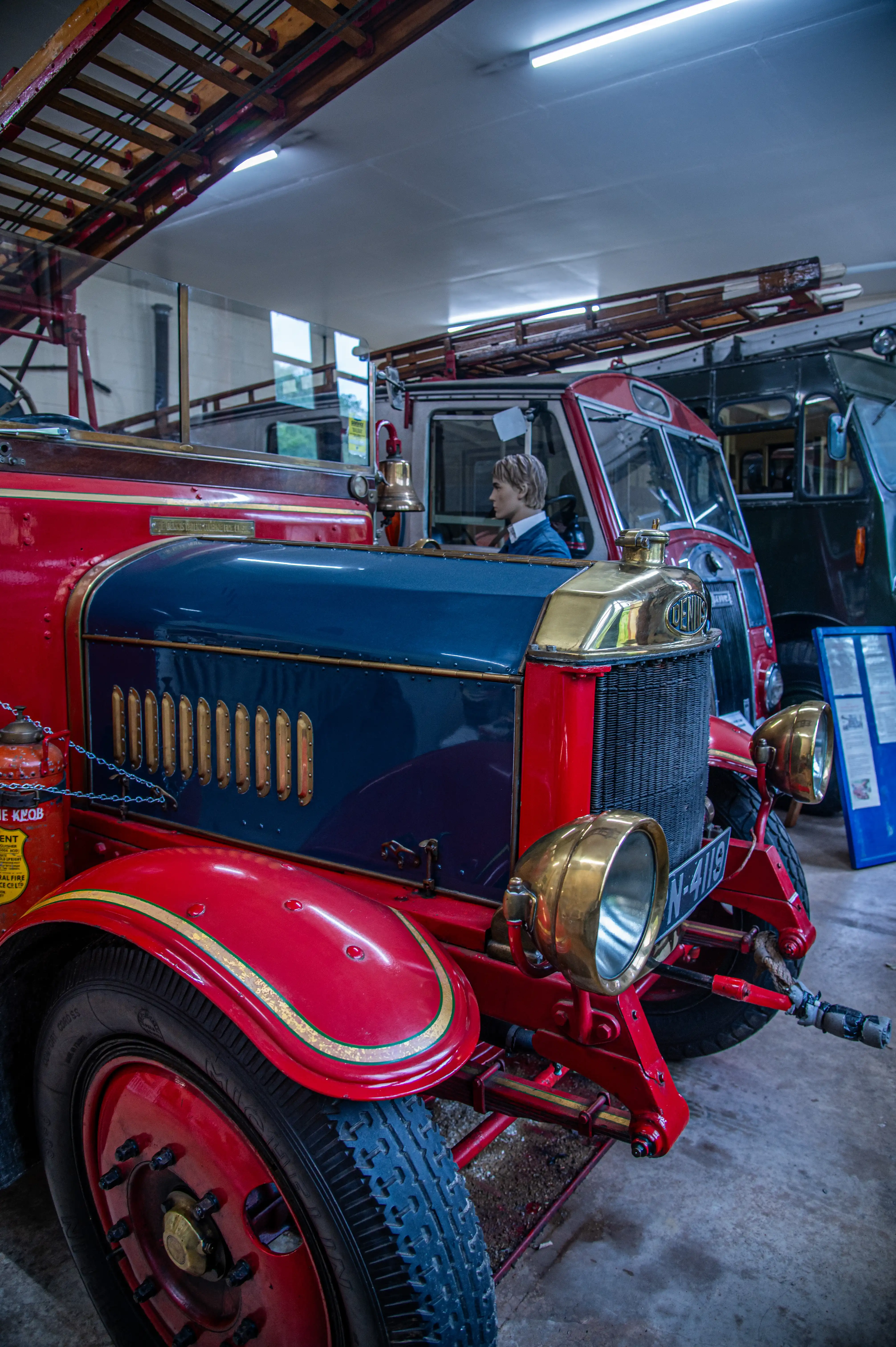 A vintage red and blue fire engine with the number 9 prominently displayed, sits in a garage. Brass headlights and accents gleam on the vehicle, which has a large hose wound around a reel on the side. Other firefighting equipment and large wooden doors with glass panes are visible in the background.