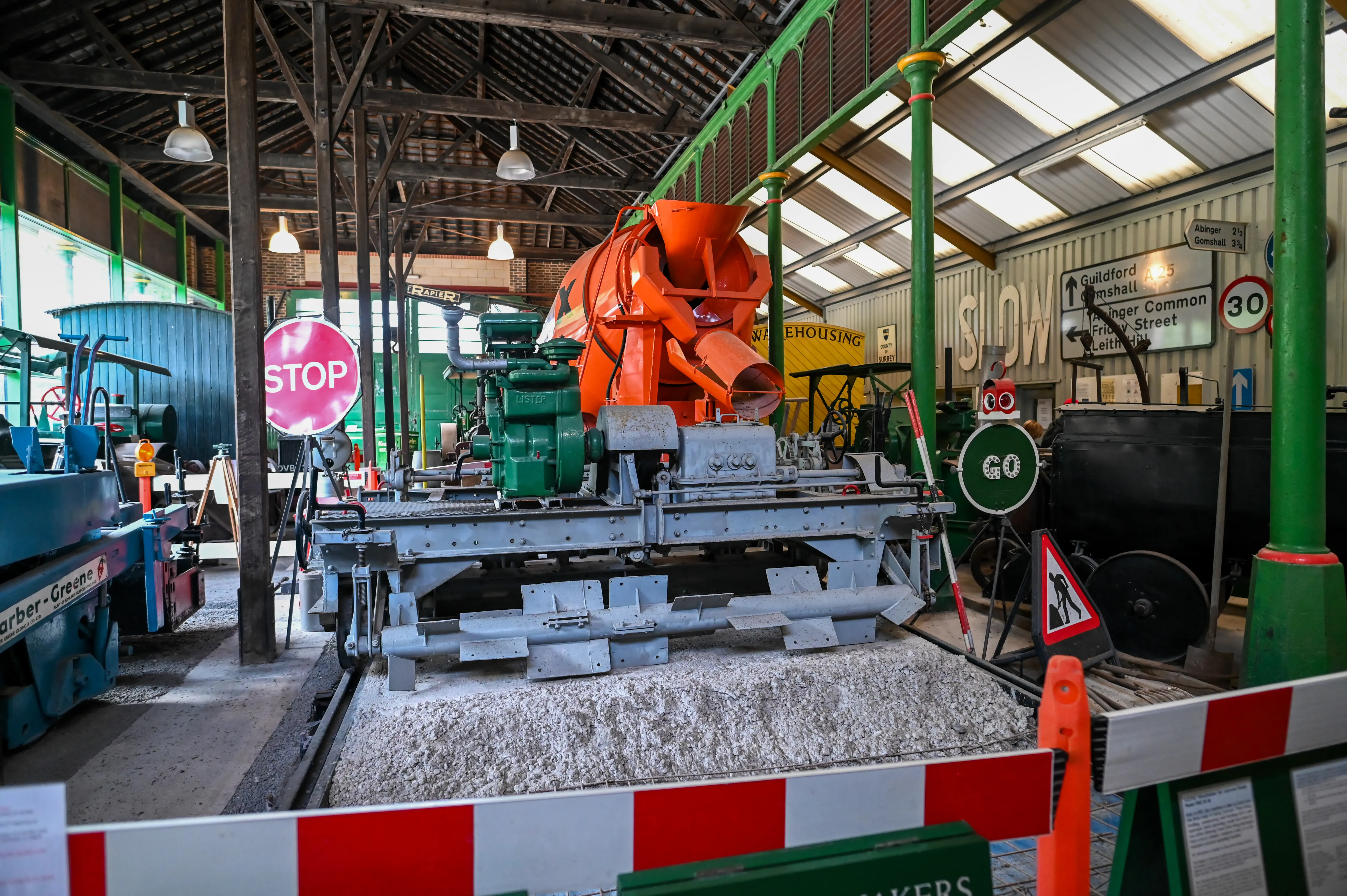 An orange concrete mixer with a green engine sits on a track in a museum. Various industrial machines, signs, and safety barriers are visible around it.