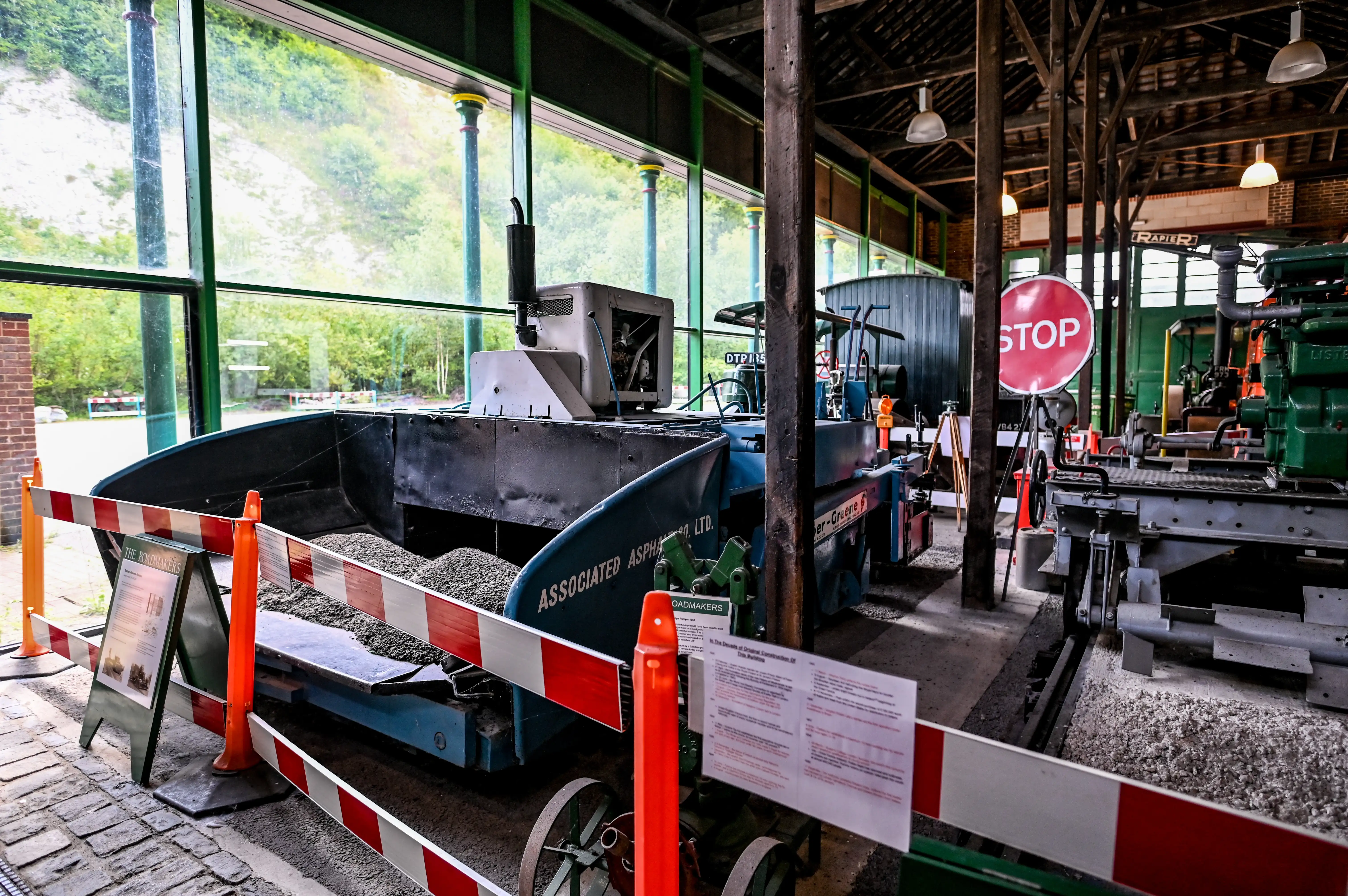 A collection of vintage road construction machines, including a large blue asphalt paving machine filled with gravel and a green engine, displayed in an indoor setting with large windows showing greenery outside. Red and white safety barriers and information signs are positioned around the machinery.