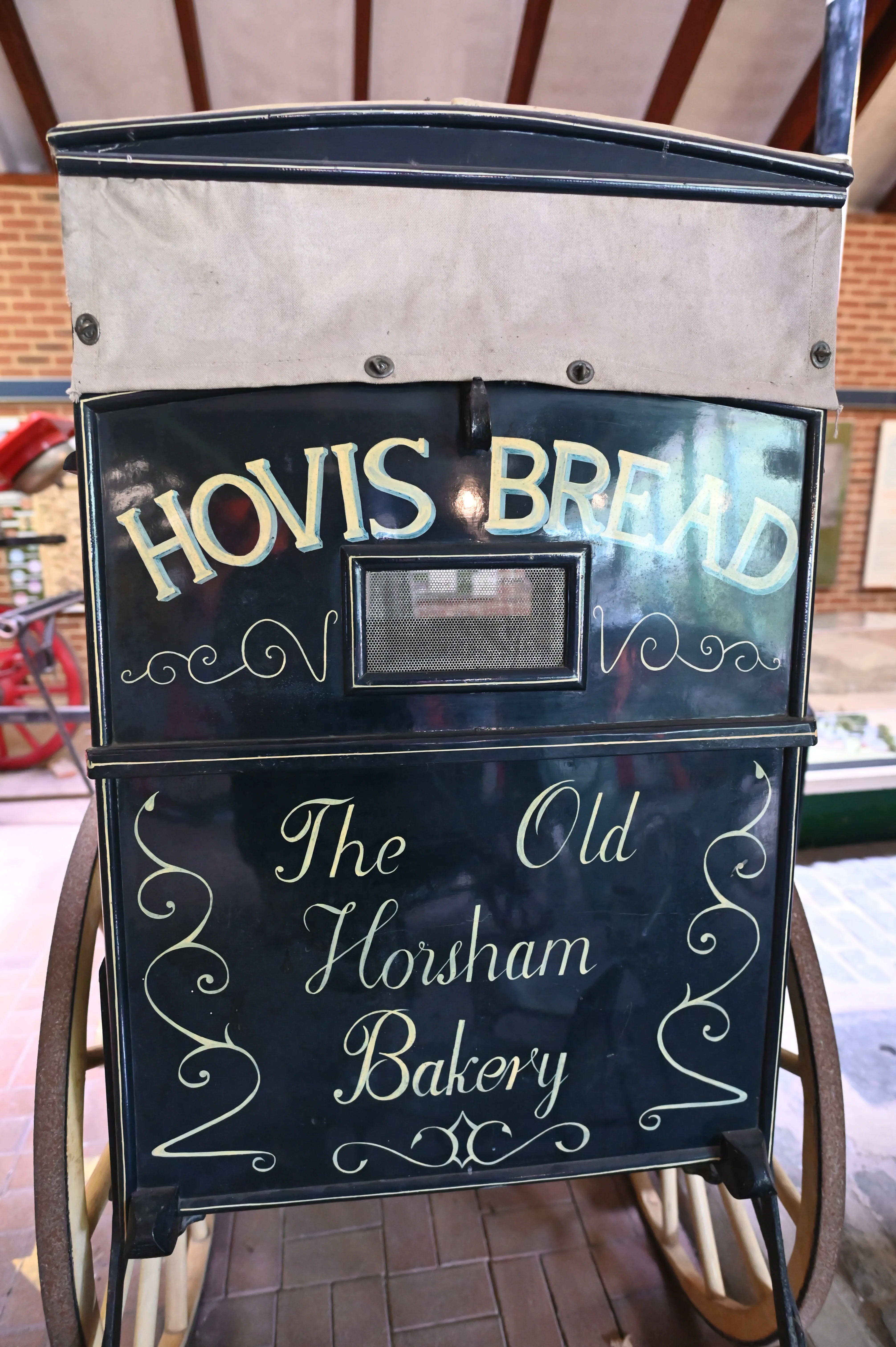 A vintage black Hovis bread delivery cart with ornate lettering. The top panel reads 'HOVIS BREAD' in gold and blue. The lower panel reads 'The Old Horsham Bakery' in elegant script.