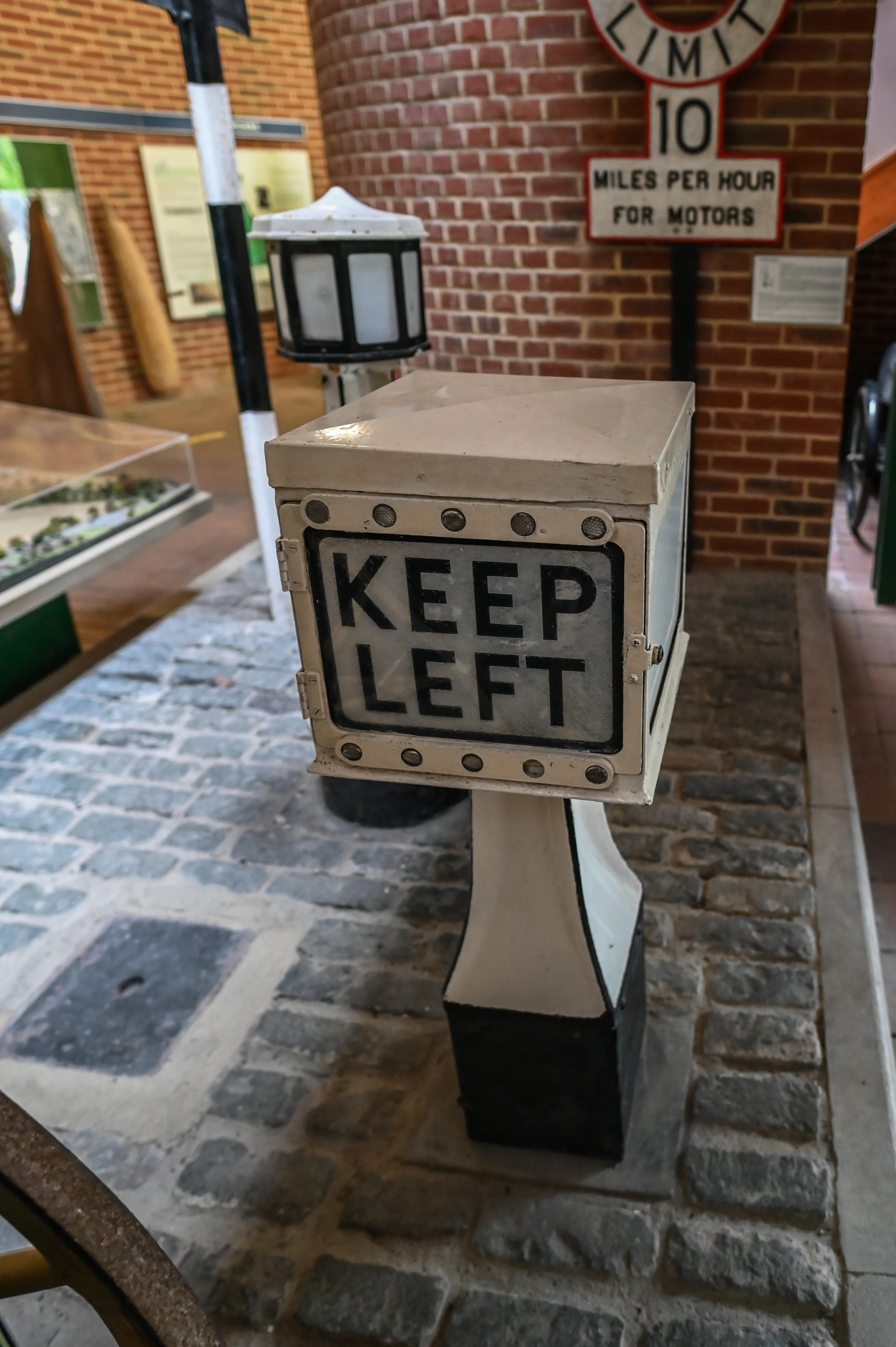 A close-up of an old white metal sign with black text reading 'KEEP LEFT'. The sign is on a pedestal and appears to be in a museum exhibit.