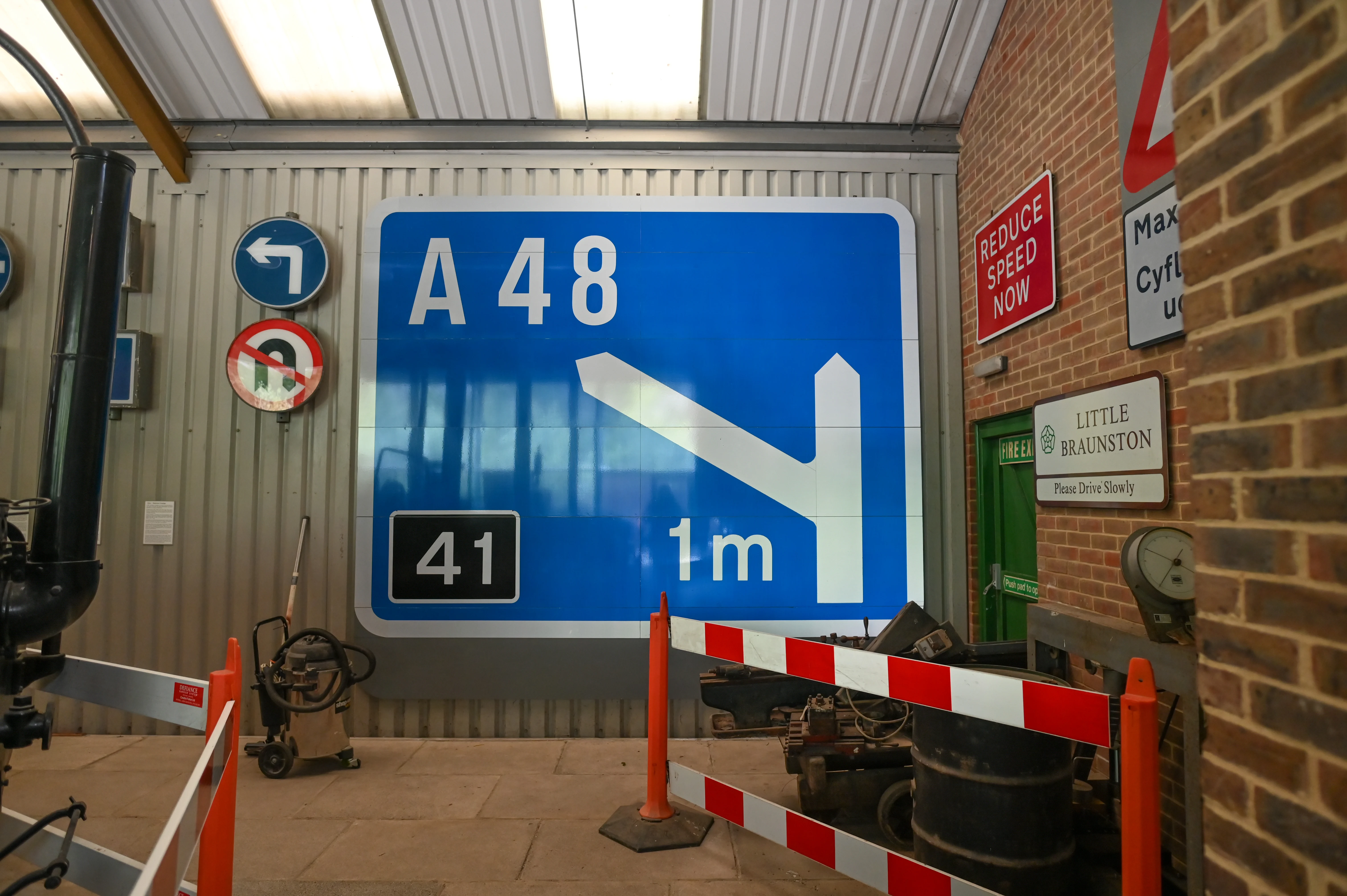A display of various British road signs mounted on a corrugated metal wall and a brick wall. The most prominent sign is a large blue directional sign for the A48, indicating junction 41 and a distance of 1 metre. Several smaller signs are also visible, along with some industrial equipment and safety barriers.