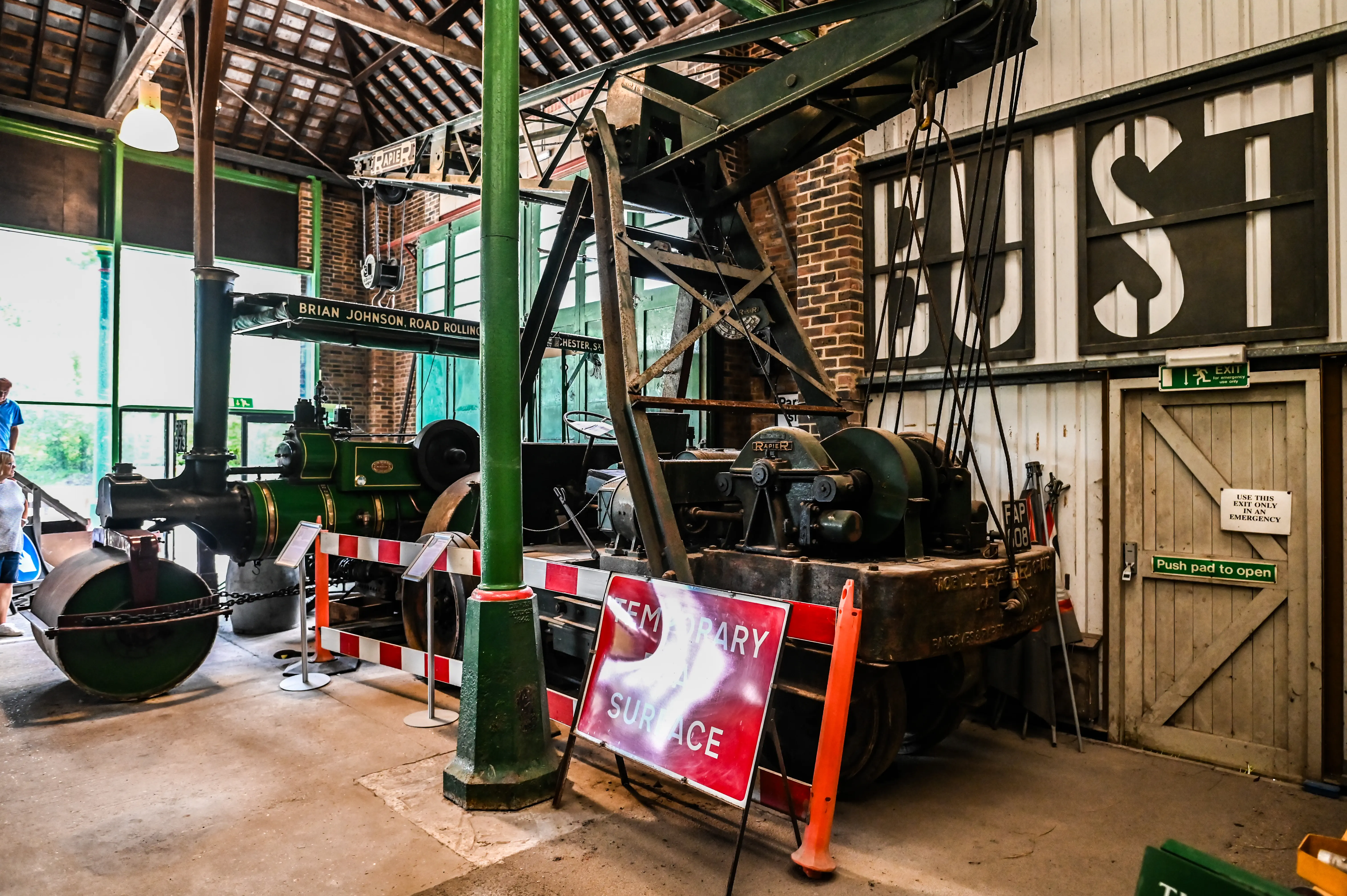 An interior view of a museum exhibit featuring a green vintage steam road roller next to a large industrial crane.