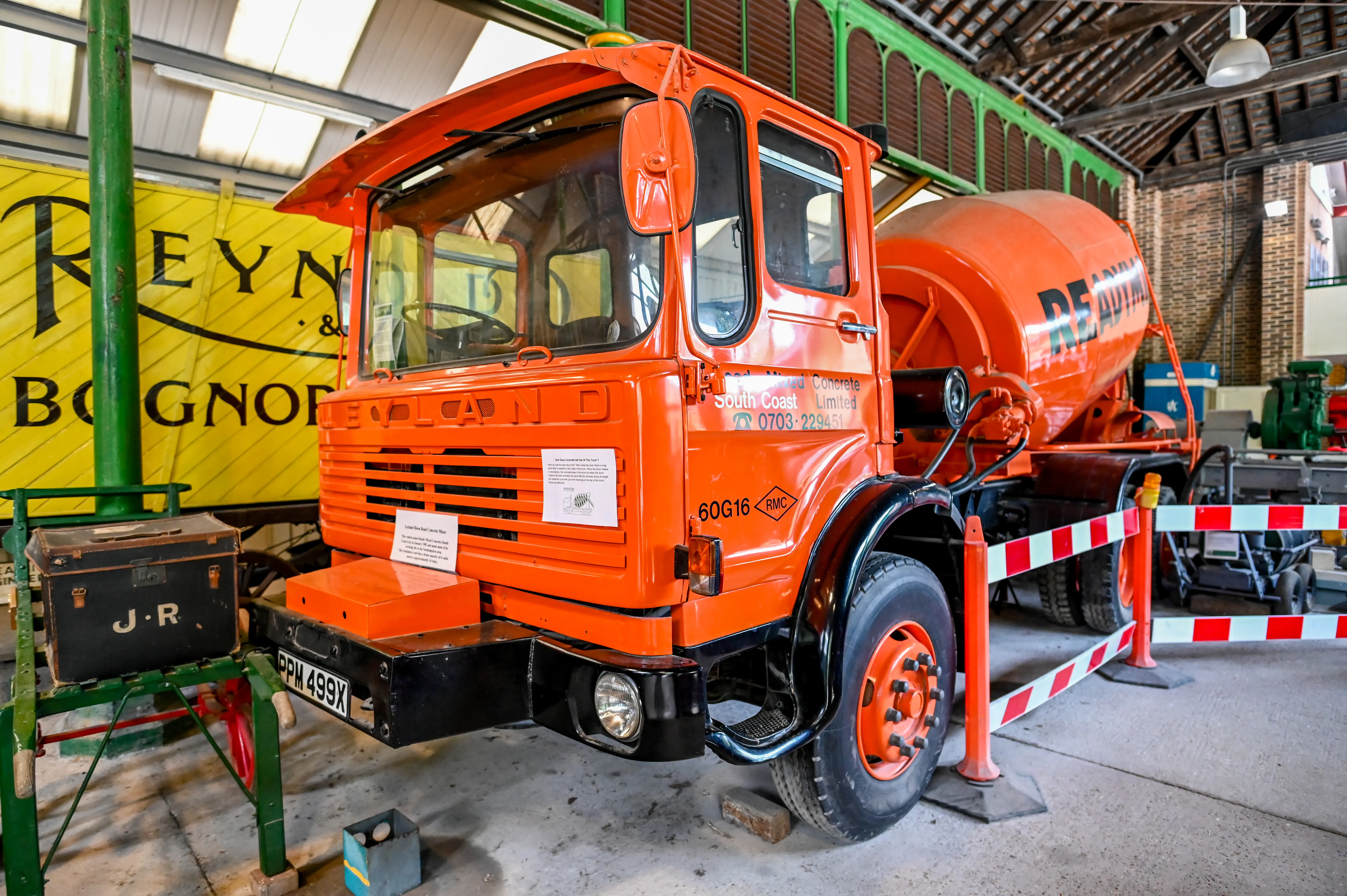 A vintage orange cement mixer truck with the registration plate PPM 499X is on display indoors. In the foreground, a dark trunk with the initials 