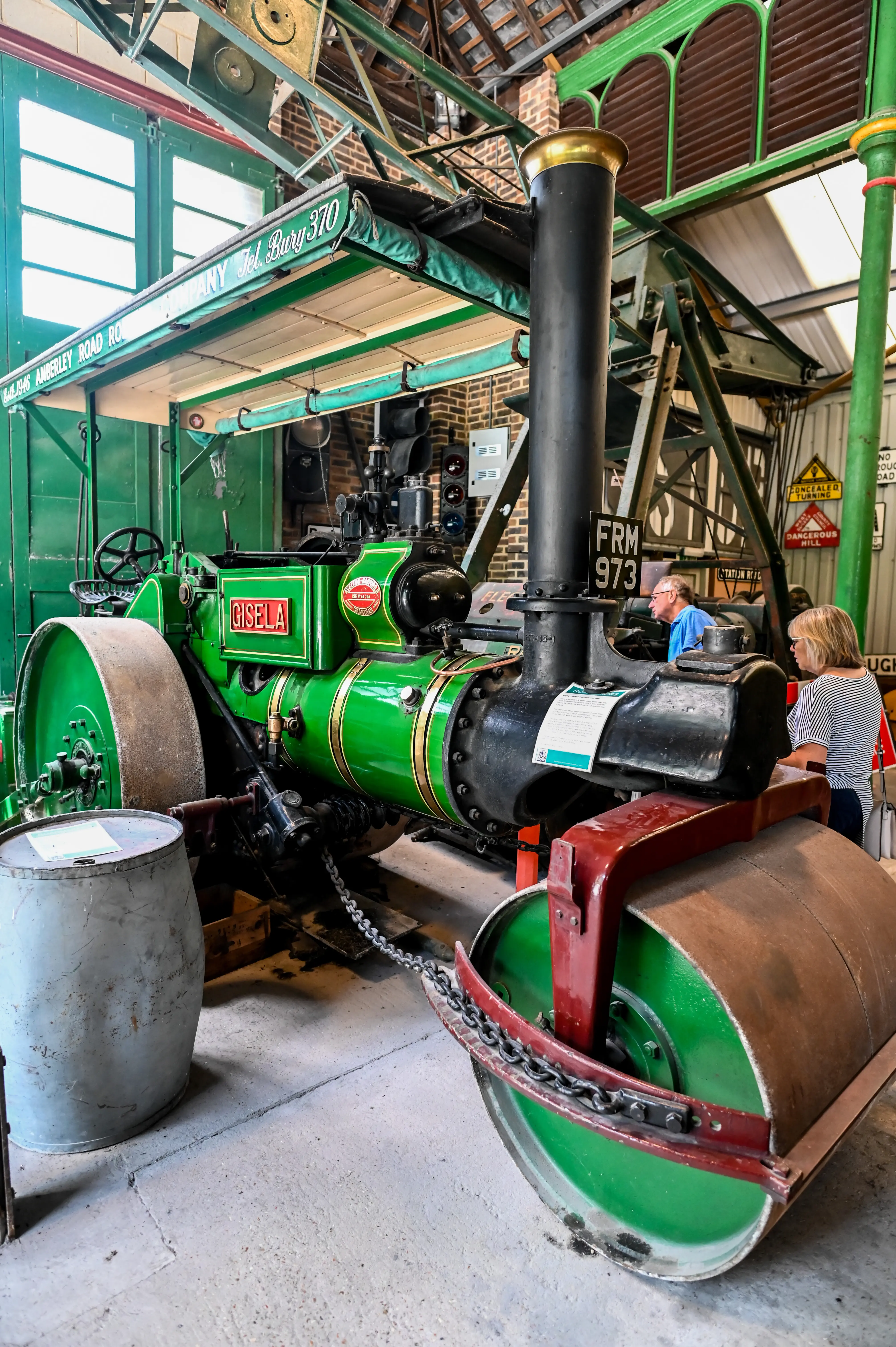 A beautifully restored vintage steam roller painted in green with red and gold accents. The steam roller is marked with 'Cisele' and 'FRM 973' and features a tall chimney, a boiler, and large metal wheels. It is displayed in a museum with industrial artifacts and visitors in the background