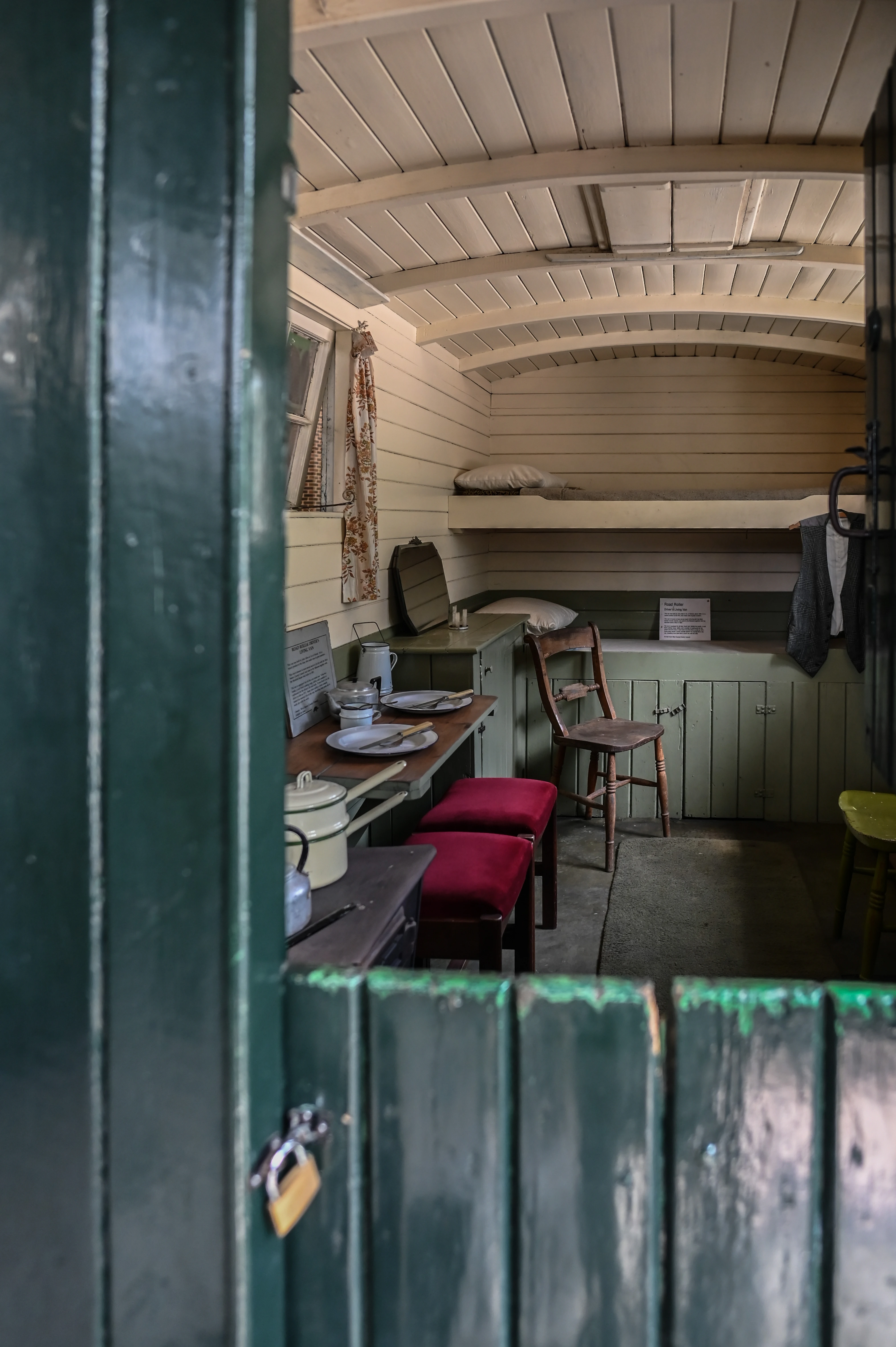 Interior view of a narrow, wooden railway carriage, showing a bunk bed above green cupboards, a table with place settings and cookware, a wooden chair, and red stools, viewed through a partly open, dark green wooden door.