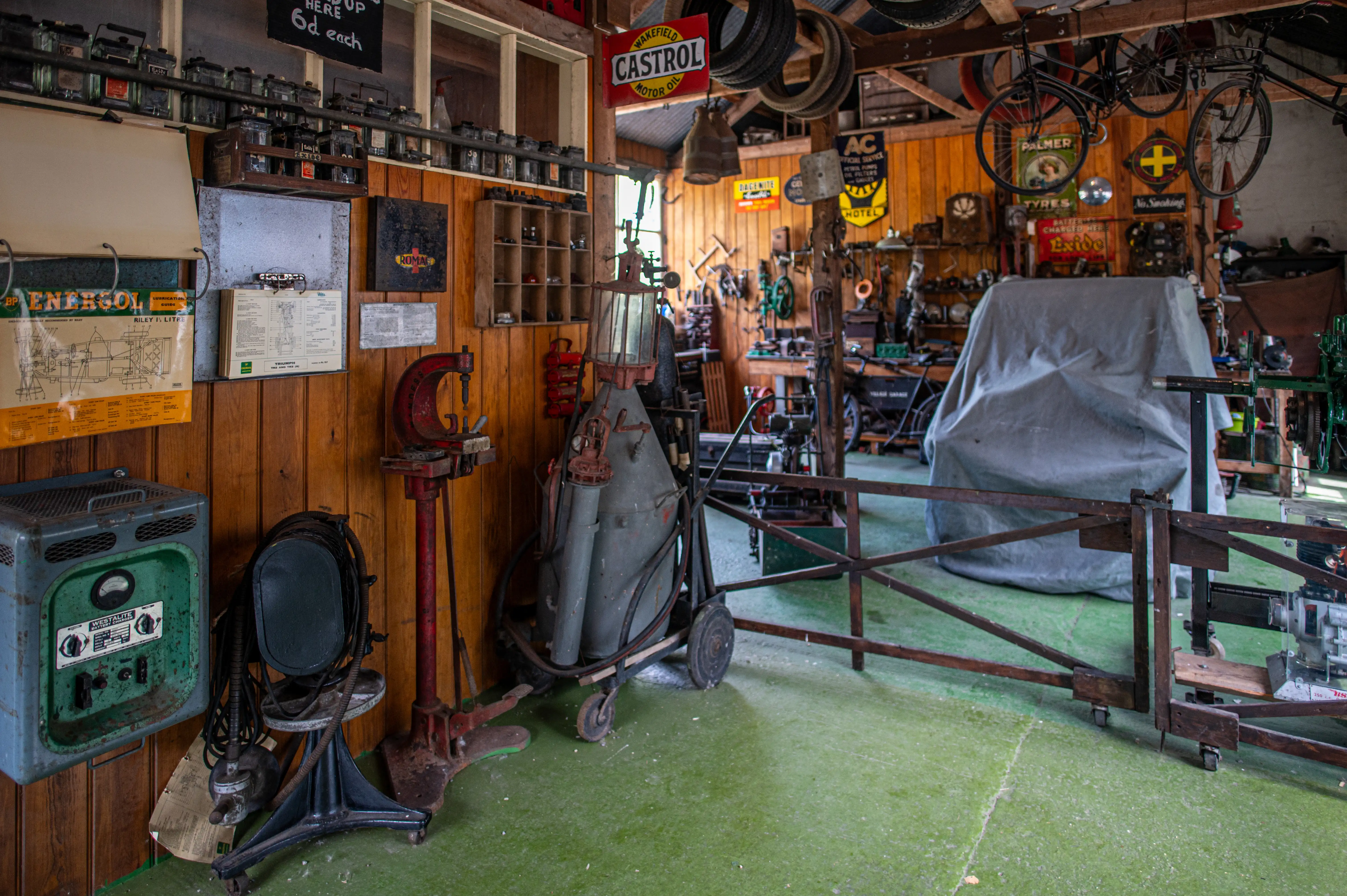 Interior view of a vintage garage workshop, filled with old tools, car parts, vintage signs, and a large object covered by a sheet.