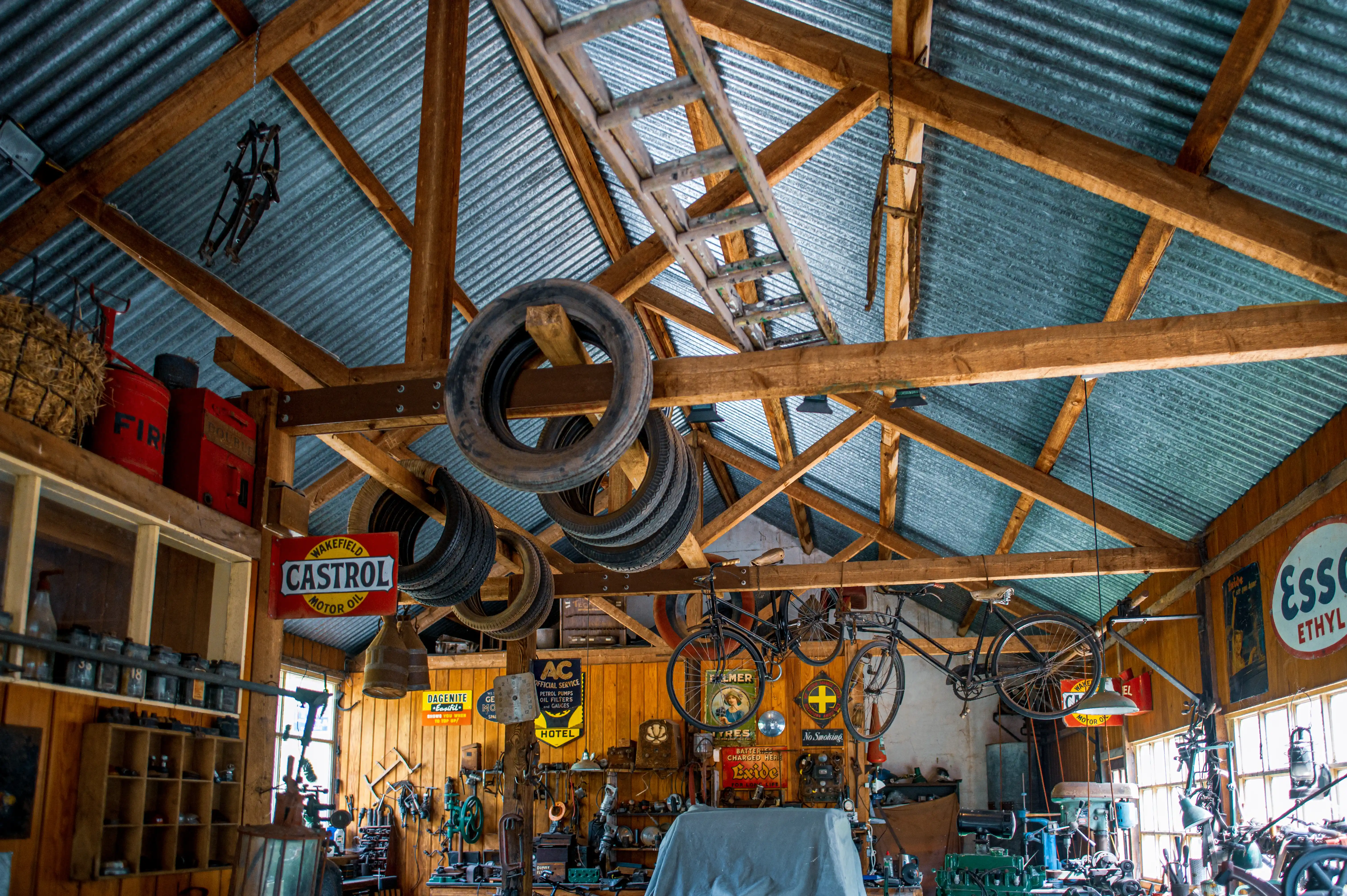 Interior view of a workshop with a corrugated metal roof, exposed wooden beams, and various vintage objects hanging and displayed, such as old tyres, bicycles, and advertising signs for Castrol and Esso.