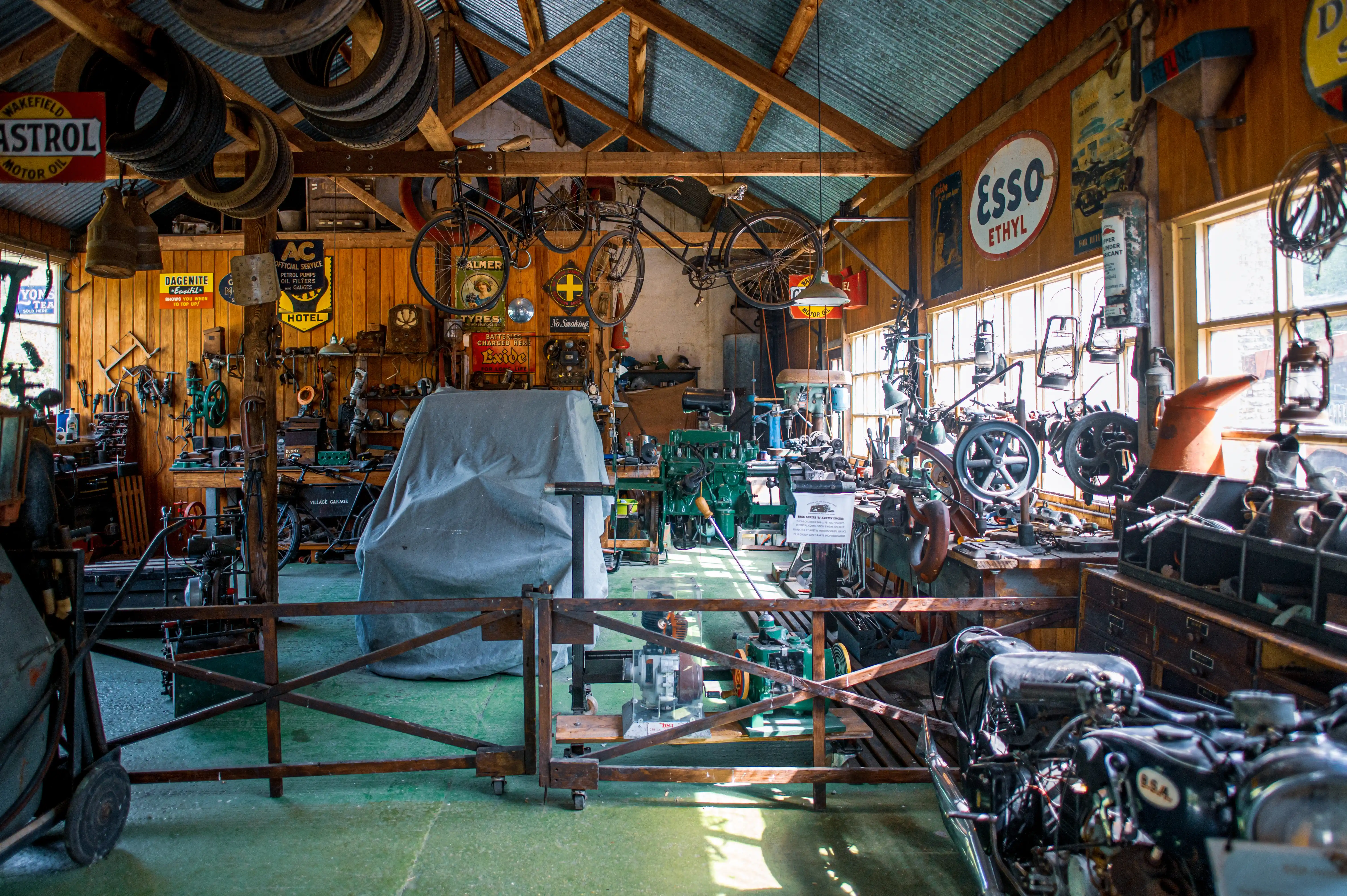 A cluttered workshop interior filled with vintage tools, machinery, bicycles, and old advertising signs. In the foreground, a motorcycle is partially visible, and a covered object sits in the centre.