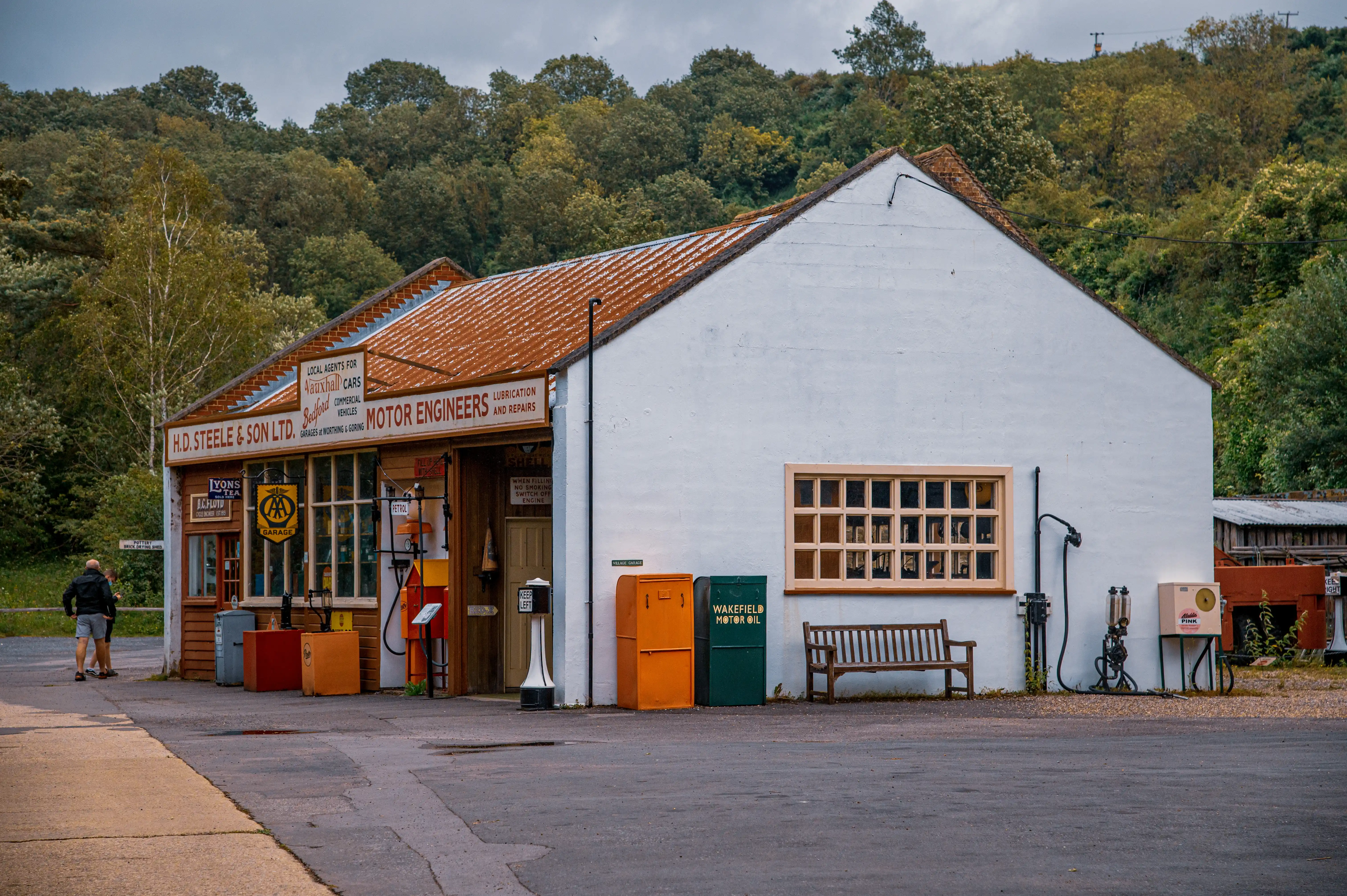 Exterior view of a white-painted, single-storey garage with a rust-coloured corrugated roof. The front of the building features traditional wooden shopfronts advertising 