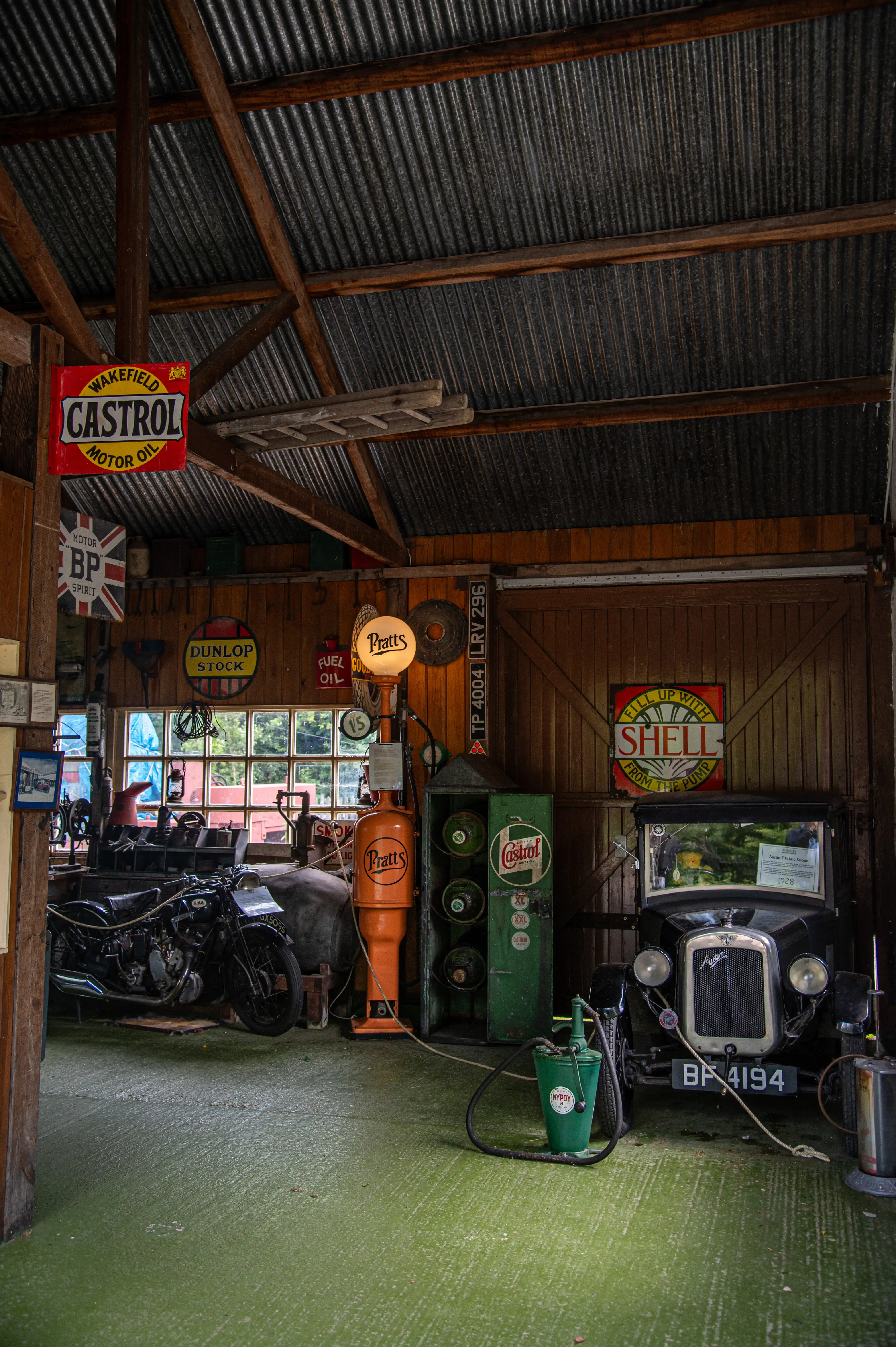Vintage garage with classic car BF 4194, motorcycle, retro fuel pumps, and motoring signs under a corrugated roof.