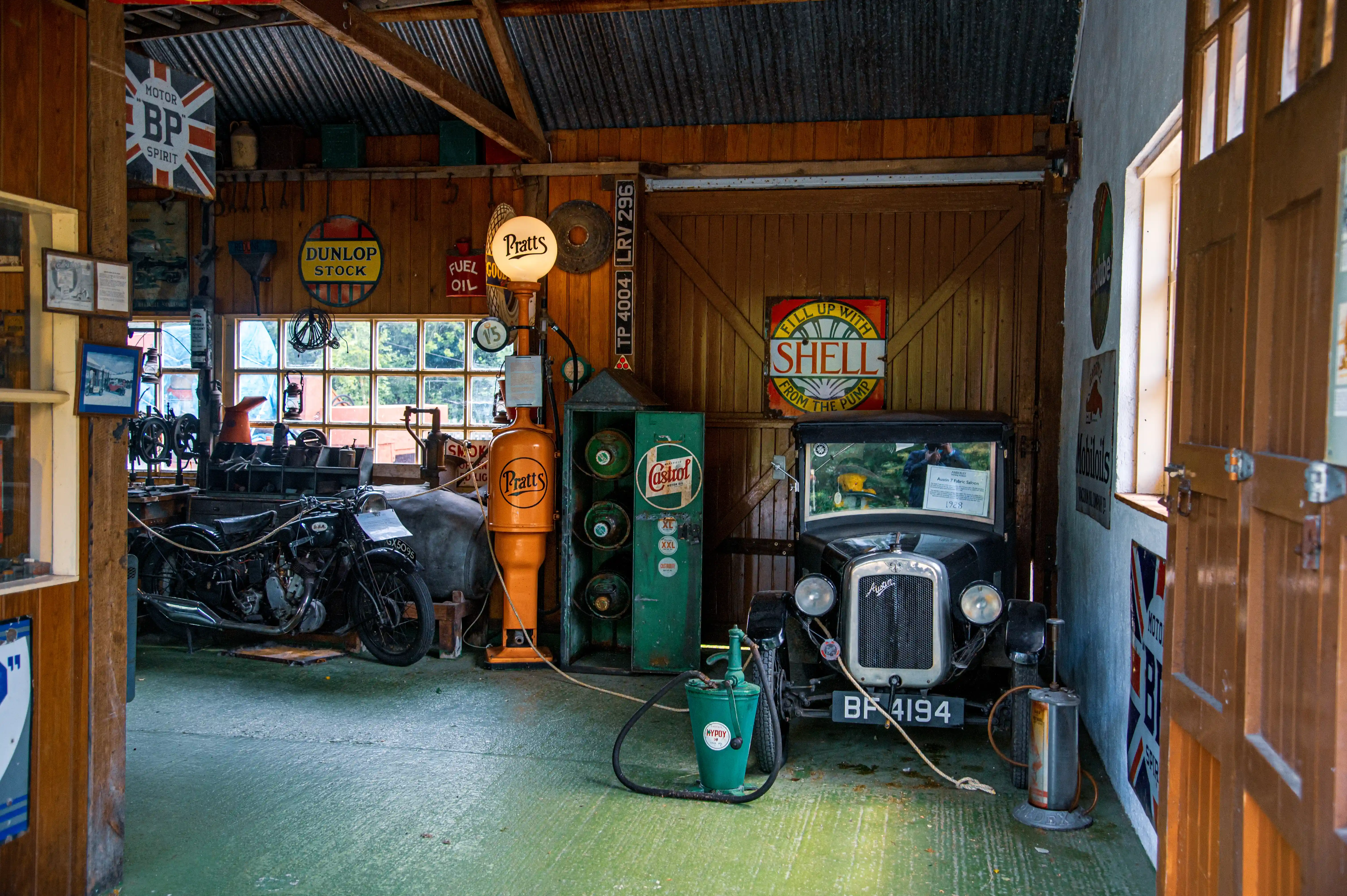 A vintage black motorcycle and a classic black Austin 7 car are parked in a wooden garage, with numerous vintage advertisements for BP, Shell, Castrol, and Dunlop displayed on the walls.