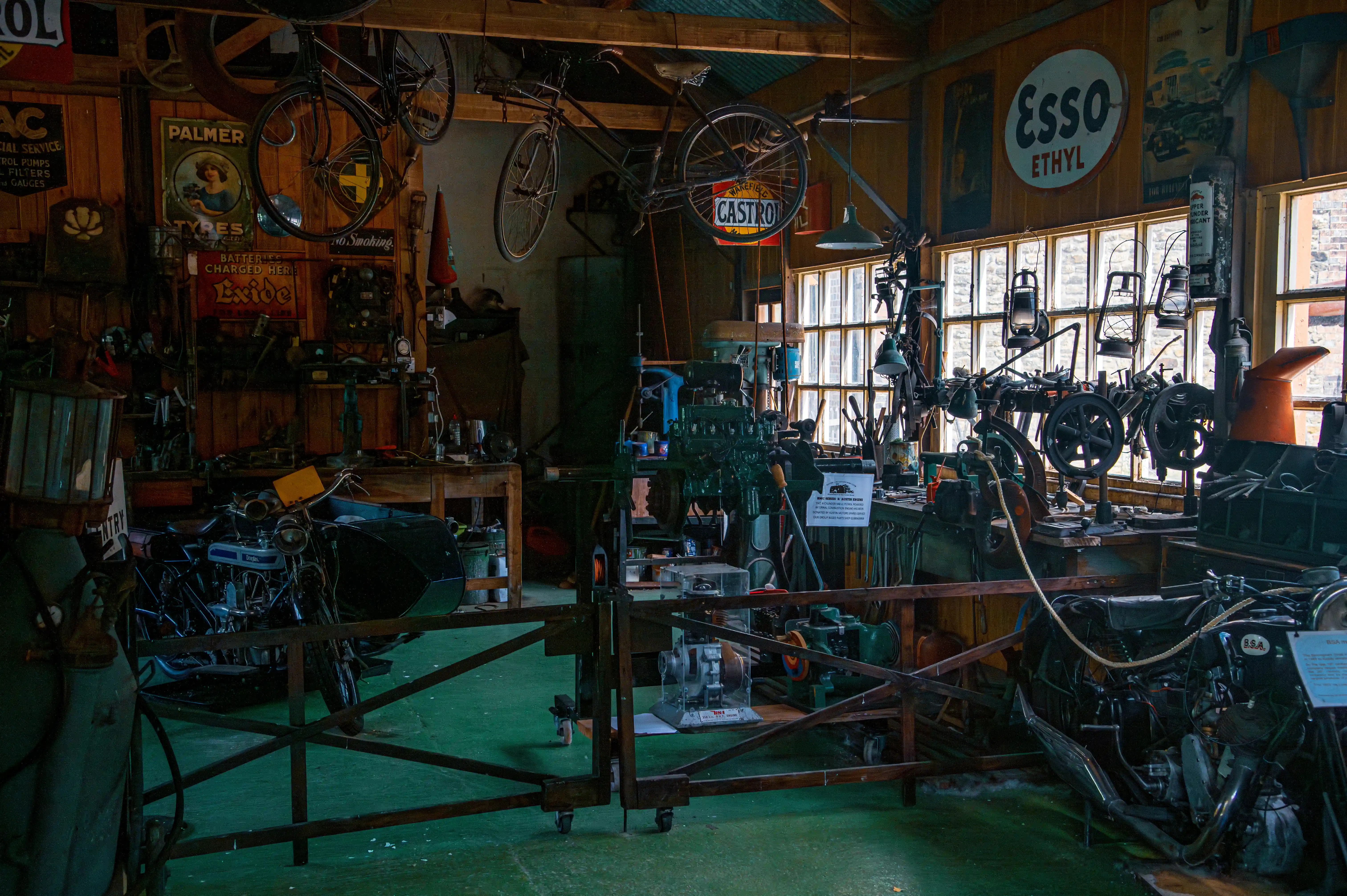 A full view of a workshop interior, with various vintage machinery, tools, bicycles hanging from the ceiling, and old signs, illuminated by natural light from large windows.
