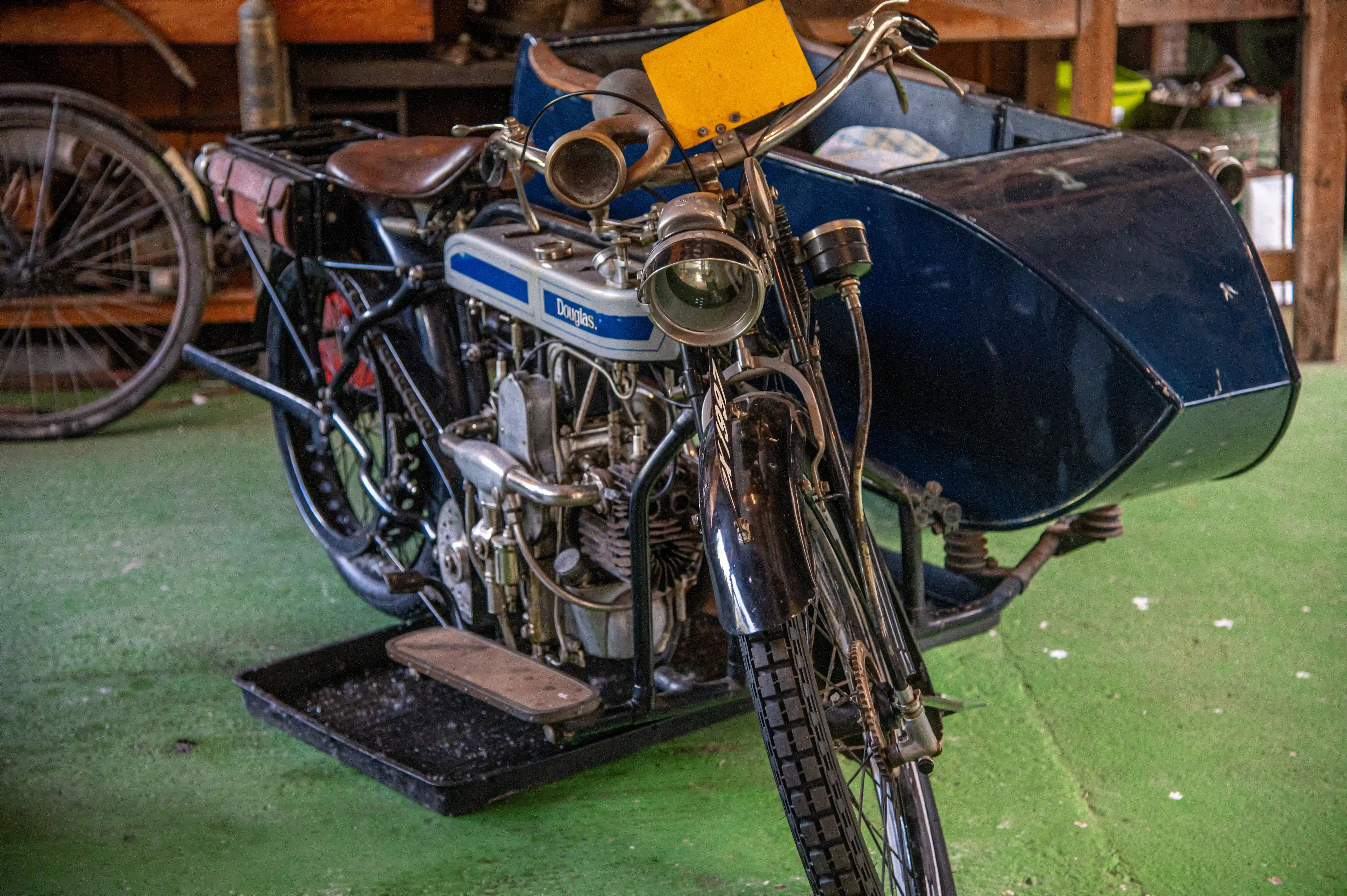 Front-three-quarter view of a vintage silver and blue Douglas motorcycle with a navy blue sidecar. The motorcycle has a large engine, chrome handlebars with a yellow sign, and a brown leather seat. A leather tool bag is on the back. The vehicle is parked on a green floor.