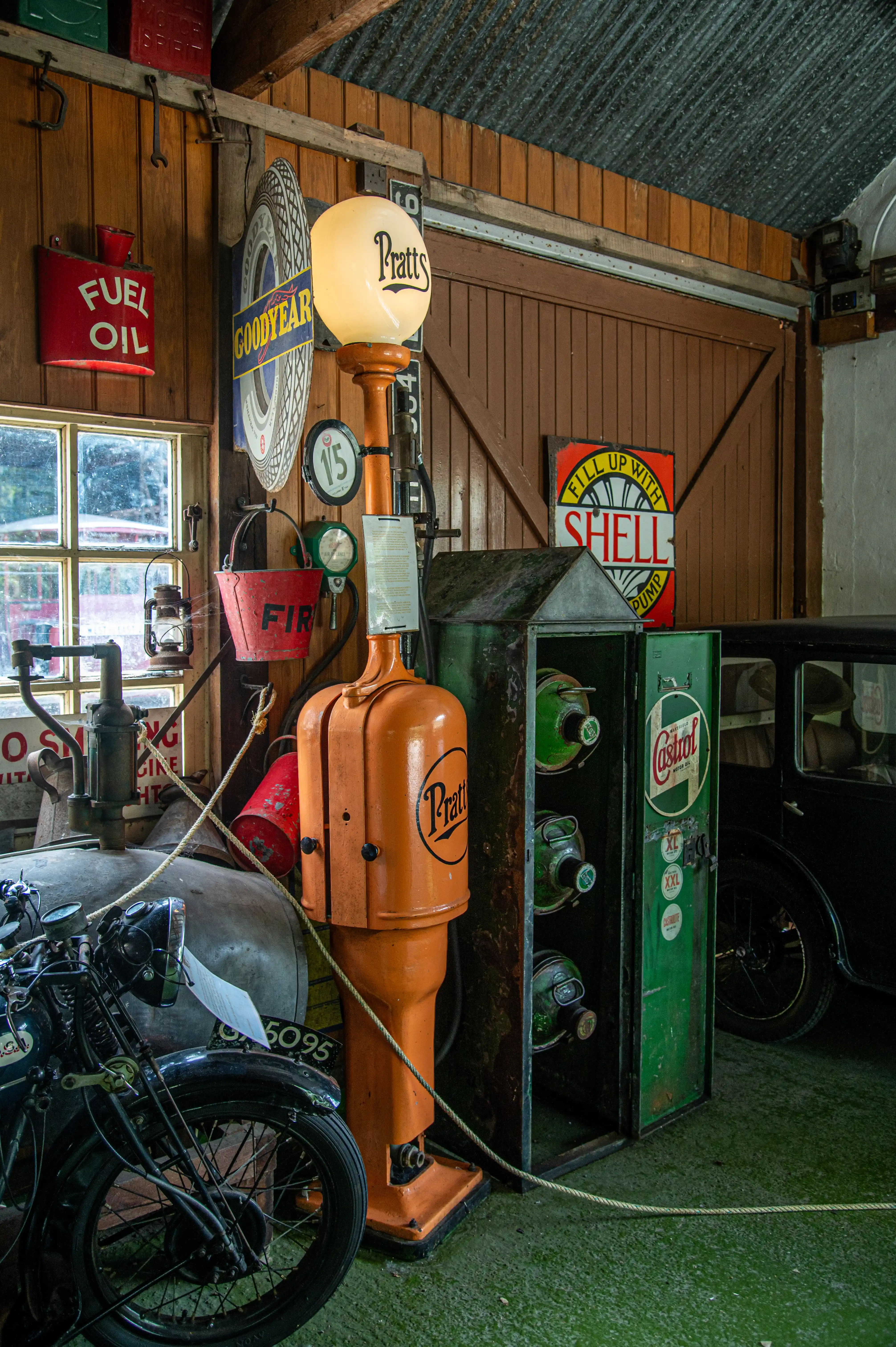 Interior of a vintage garage with an orange Pratt's petrol pump and a green Castrol oil cabinet. A classic motorcycle is parked to the left, and vintage signs for Fuel Oil, Goodyear, and Shell are visible on the wooden walls.