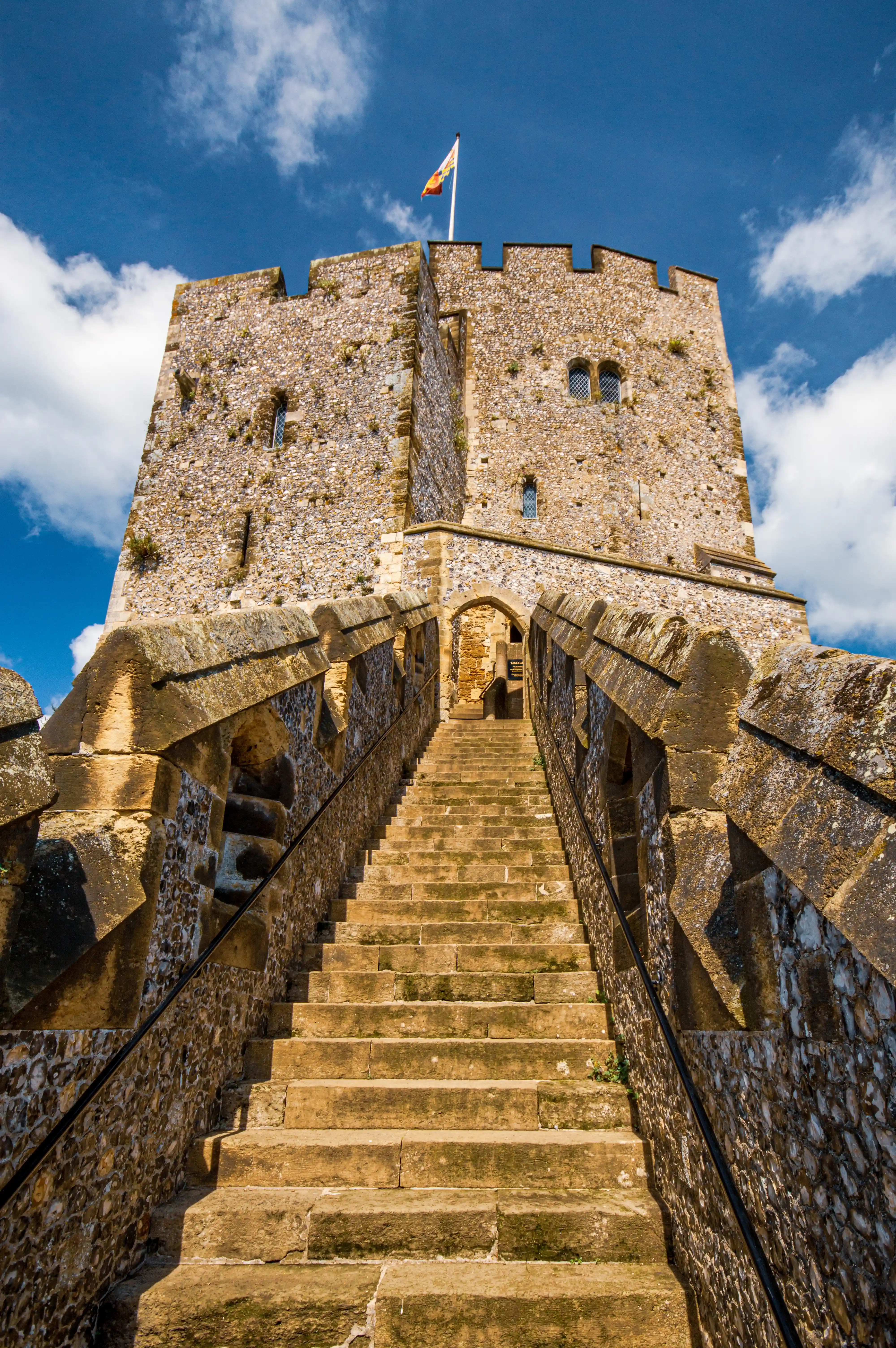 A wide stone staircase with battlements on either side leads upwards towards a medieval stone castle keep under a bright blue sky with white clouds. A flag flies from the top of the keep.