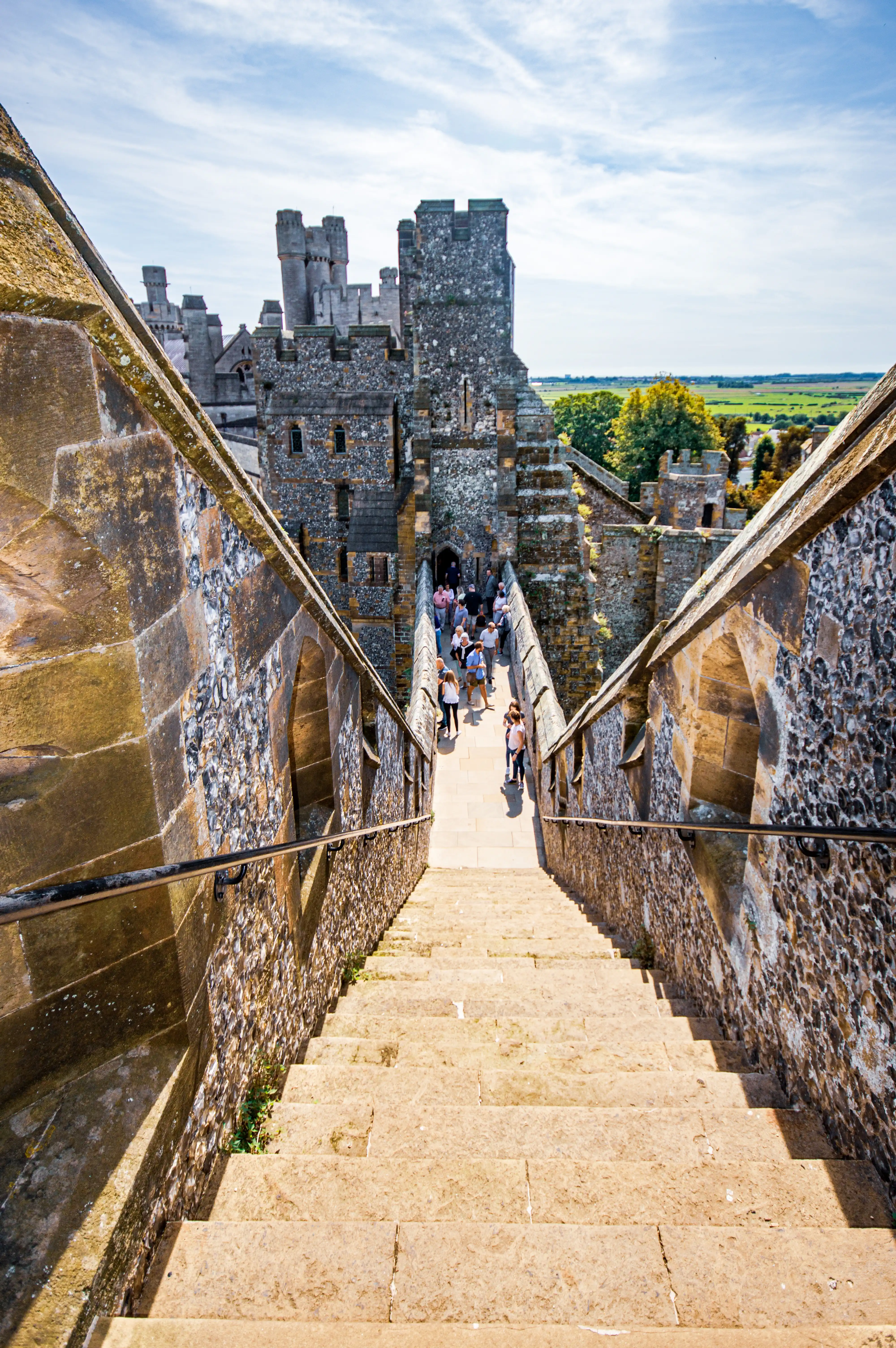 Stone staircase leading down into a medieval castle courtyard with towers, battlements and visitors, set against a rural landscape under a partly cloudy sky.