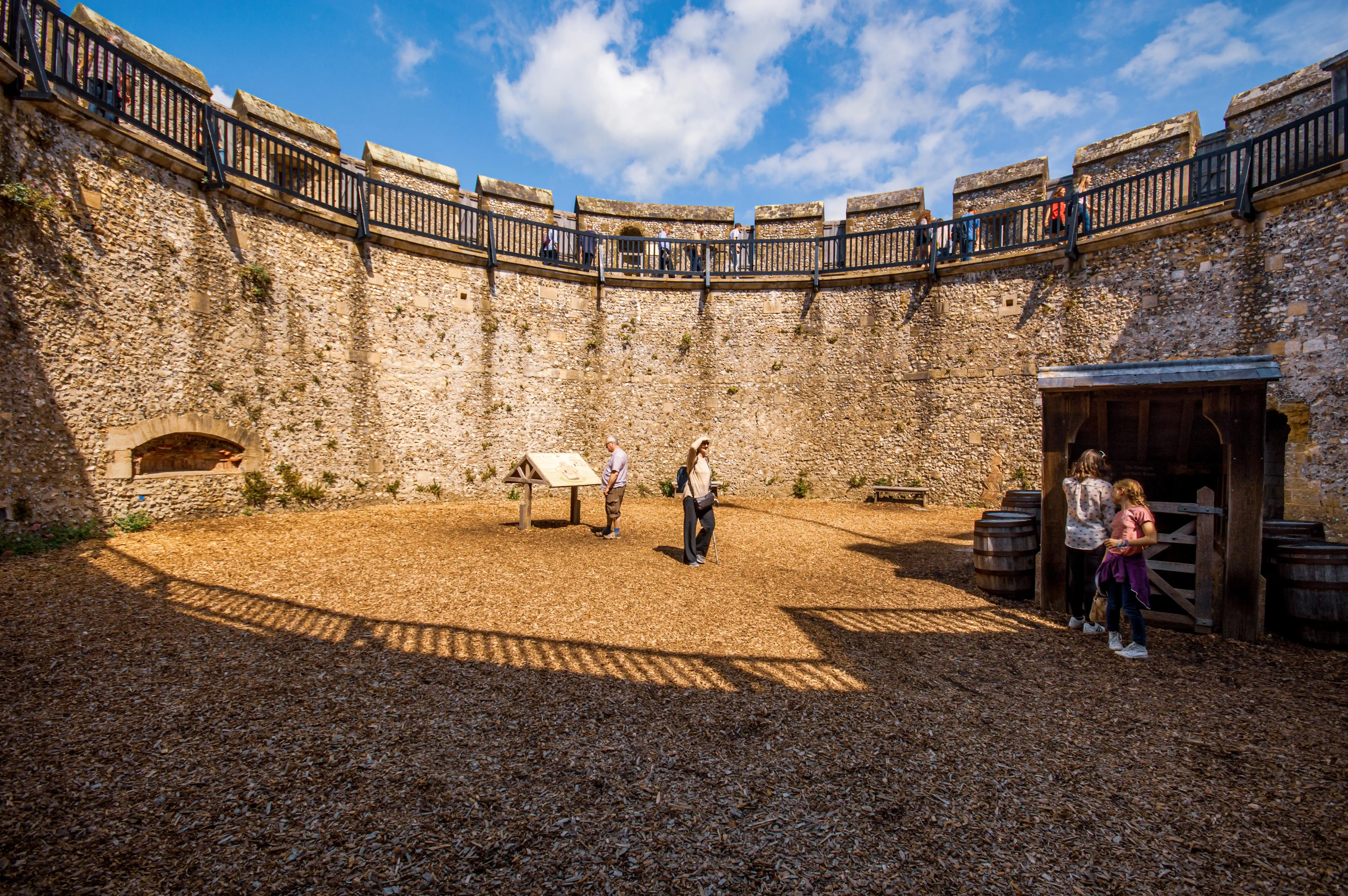 A view looking into the courtyard of a historic stone castle. People are visible on the battlements above and in the courtyard below, near an information board and a small wooden building with barrels.