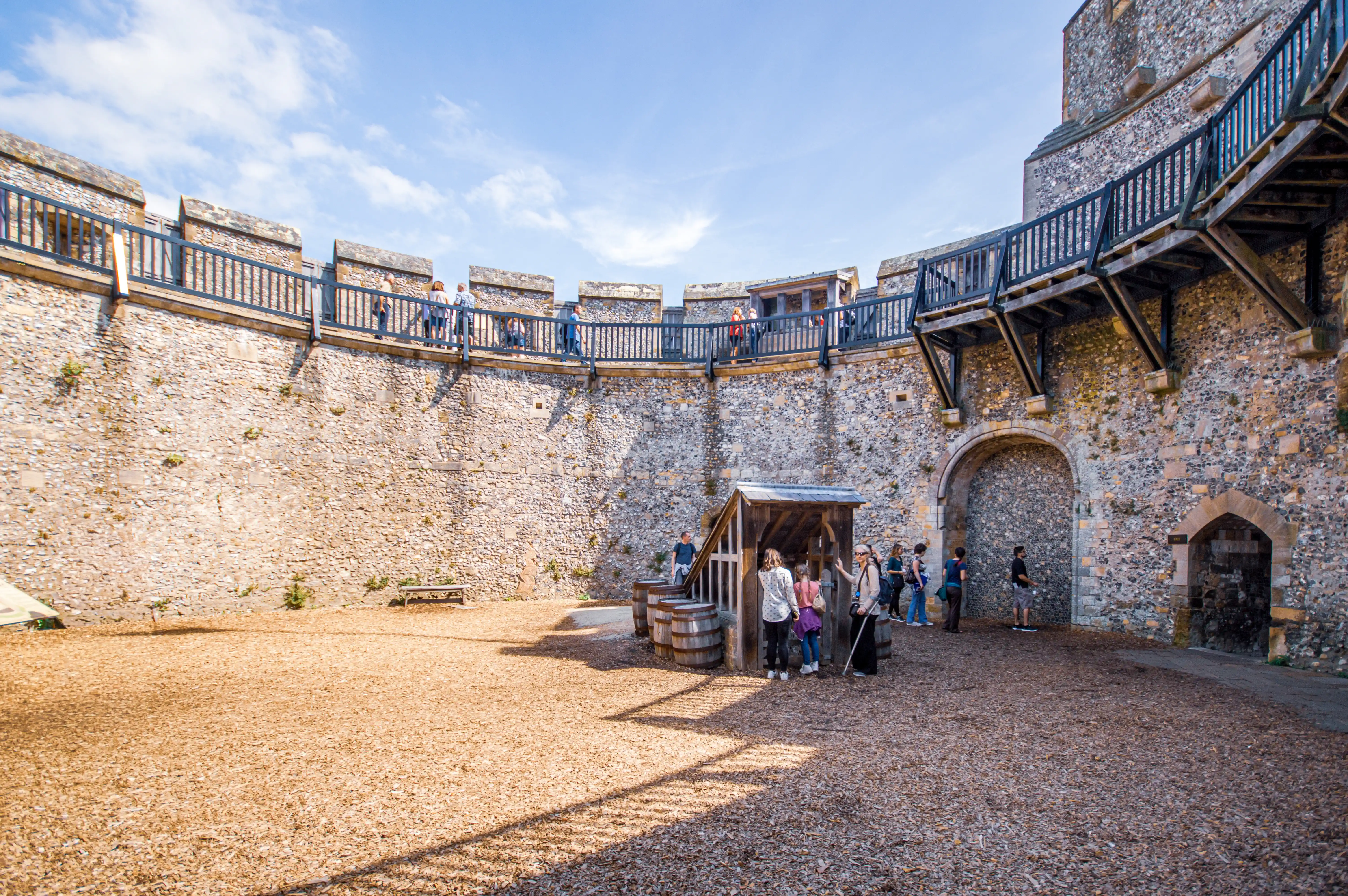Historic stone castle courtyard with tall walls, a wooden upper walkway and visitors gathered around a small timber shelter.