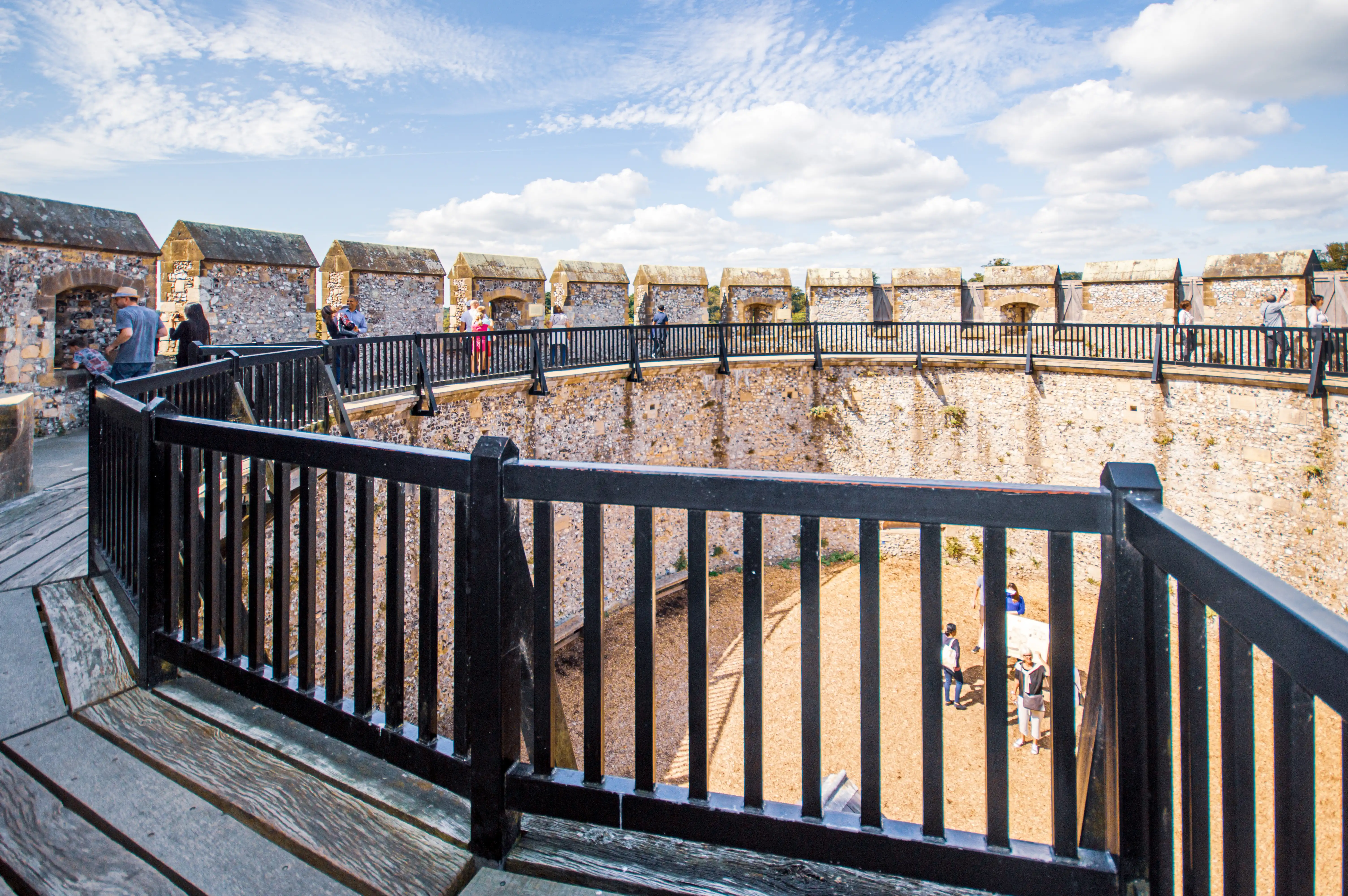 A view from a wooden walkway overlooking a historic castle's stone walls and courtyard. People are visible walking along the ramparts under a blue sky with fluffy white clouds.
