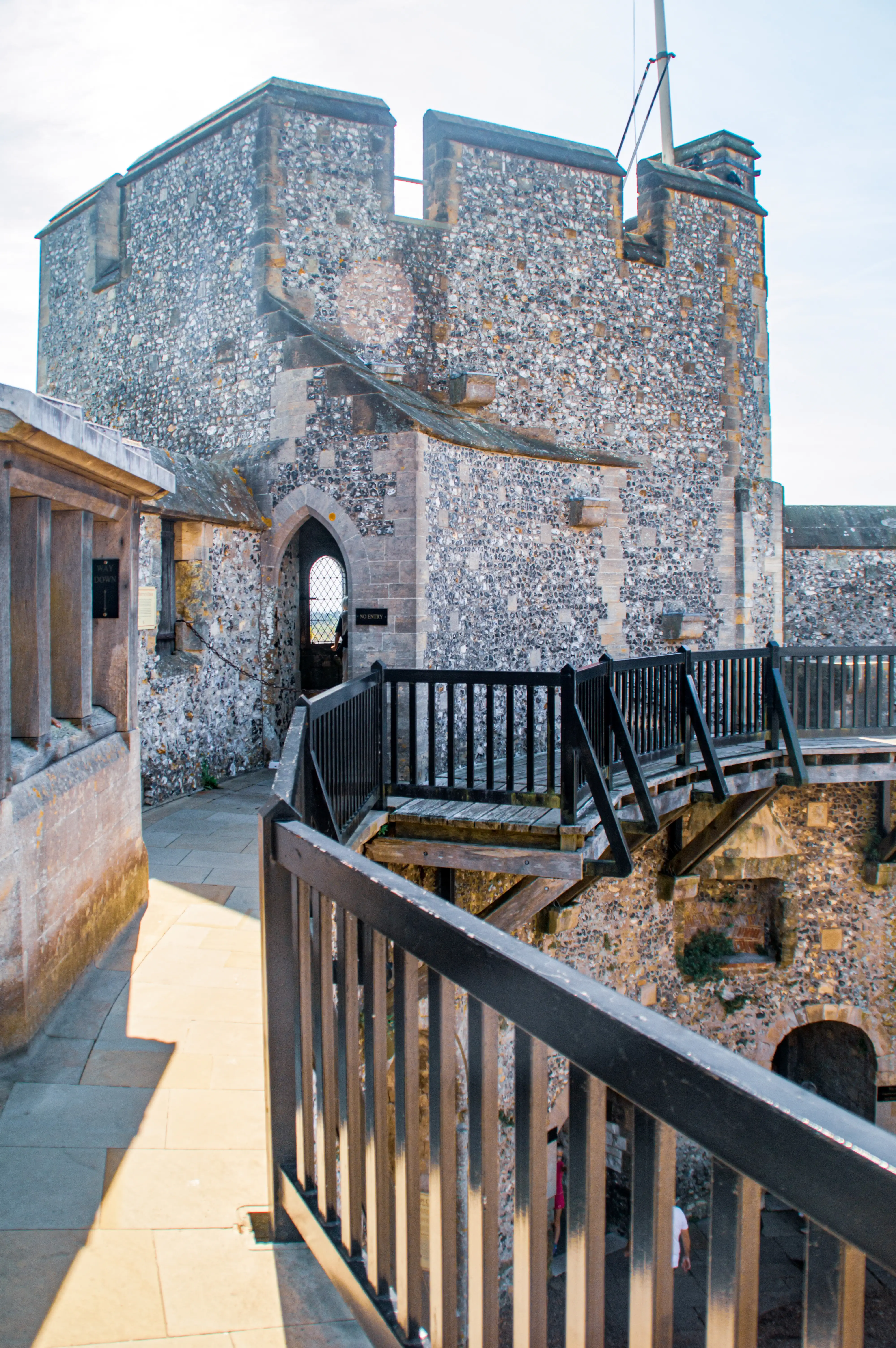 A wooden footbridge with black railings curves towards the entrance of a stone castle, which features crenellated battlements and an arched doorway.