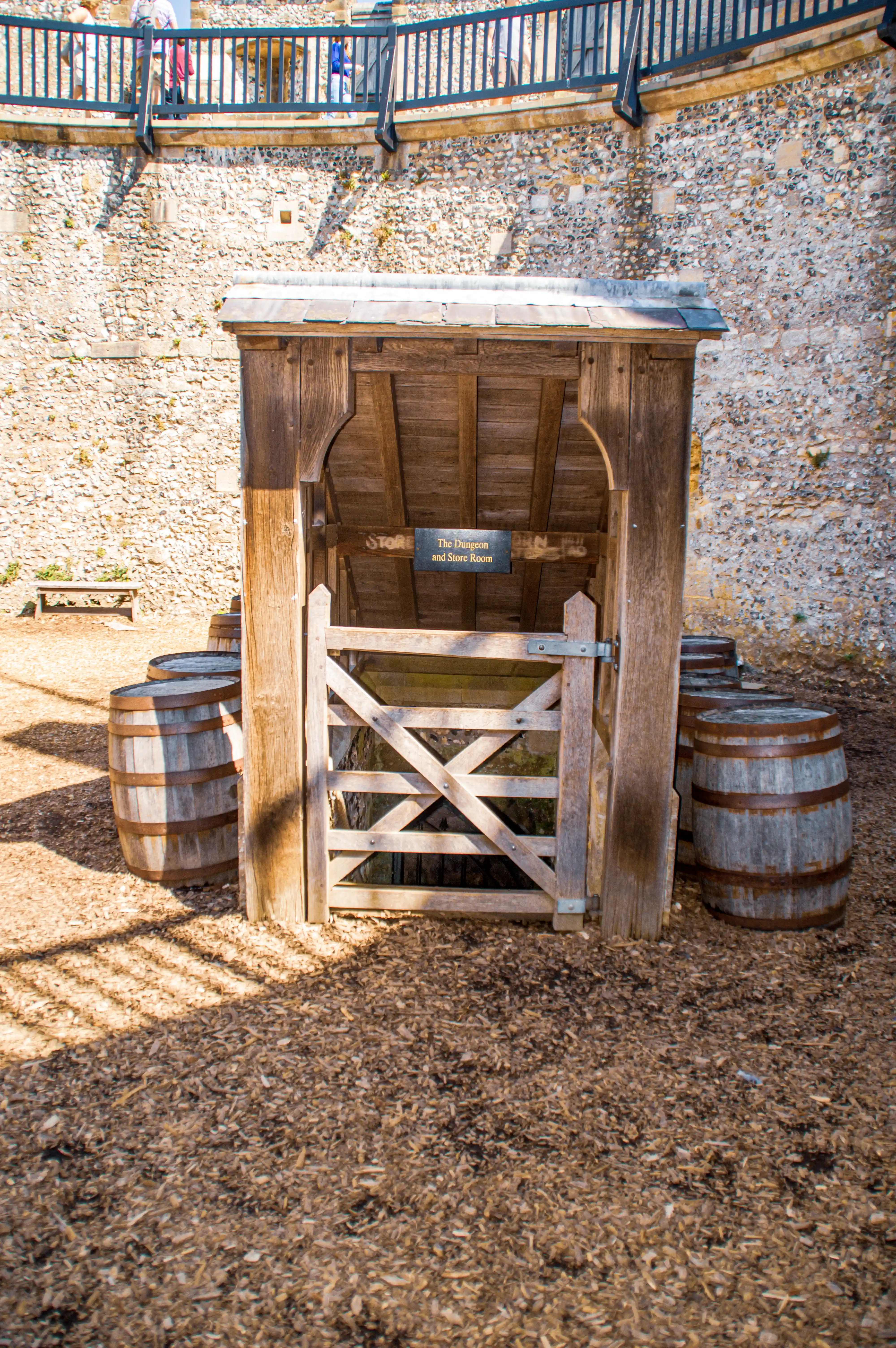 A rustic wooden gate and doorway, labelled 'The Dungeon and Store Room,' stands in front of a historical stone wall. Several weathered wooden barrels are placed around the entrance, and a wooden walkway with people can be seen above.