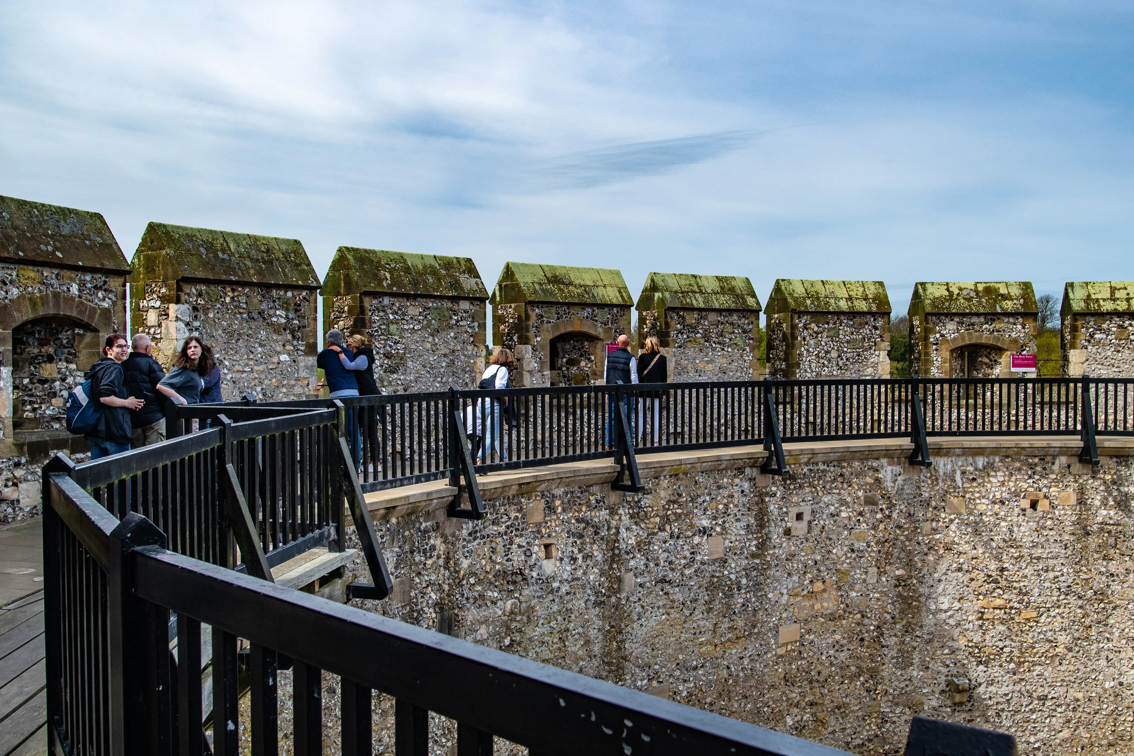 People walking on a stone walkway atop a castle wall with a series of small stone shelters and a black metal railing.
